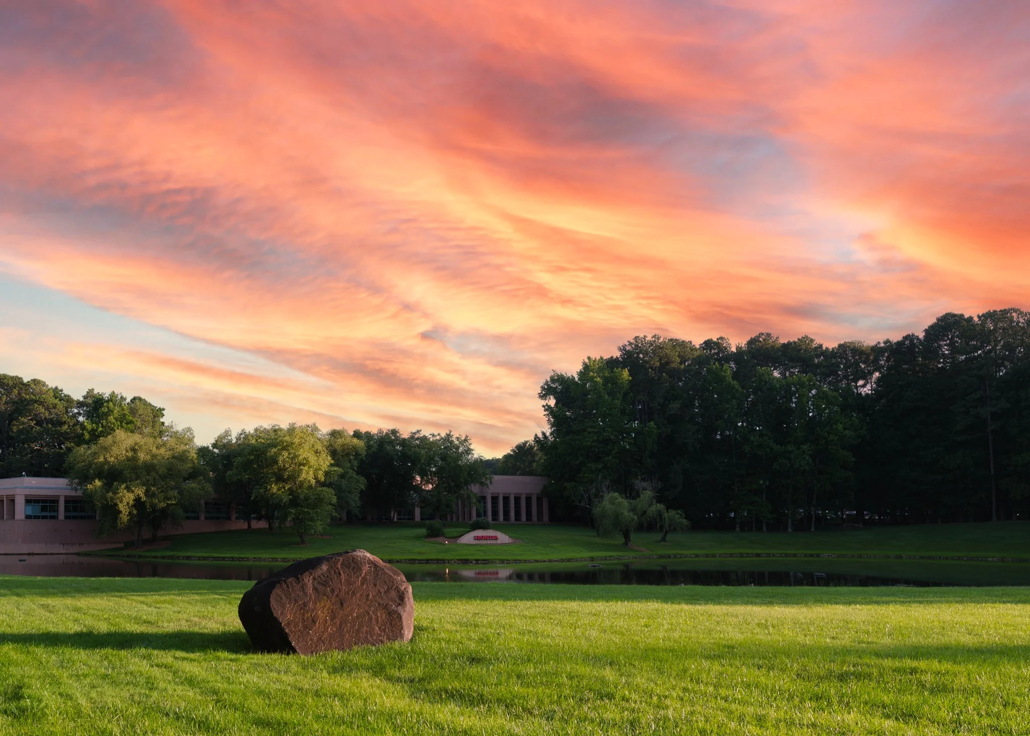 A peaceful park scene at sunset with a pink and orange sky, green grass, a large rock in the foreground, a pond, and trees. | Real Estate Photography | Alpharetta | Milton | Buckhead | Atlanta | Drone | American Honda Finance Company | Architecture p