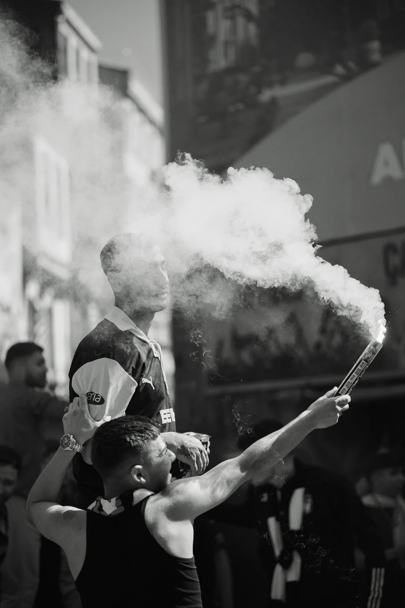 A young man is standing on a sports field, blowing a thick cloud of vape vape while holding a vape vape device. Other people are visible in the background.