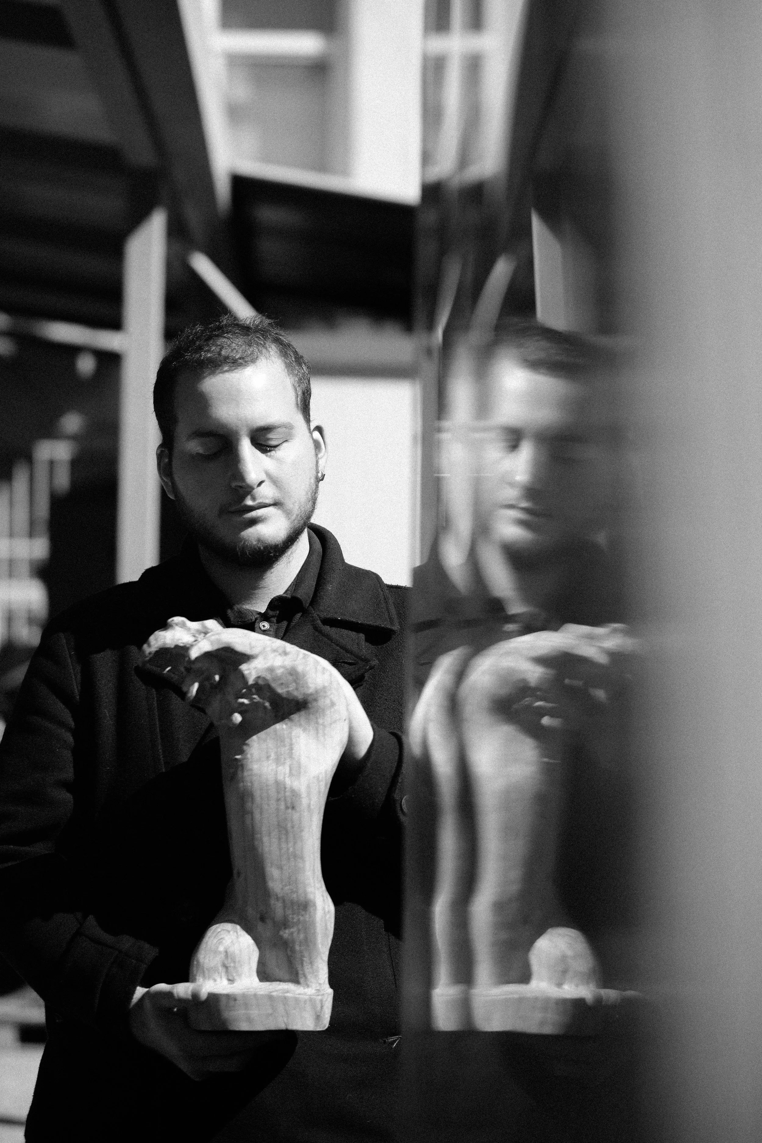 A person holding a large ceramic or porcelain sculpture of a dog with a fluffy tail, seen in front of a mirror which reflects their face and the sculpture in a black-and-white photograph.