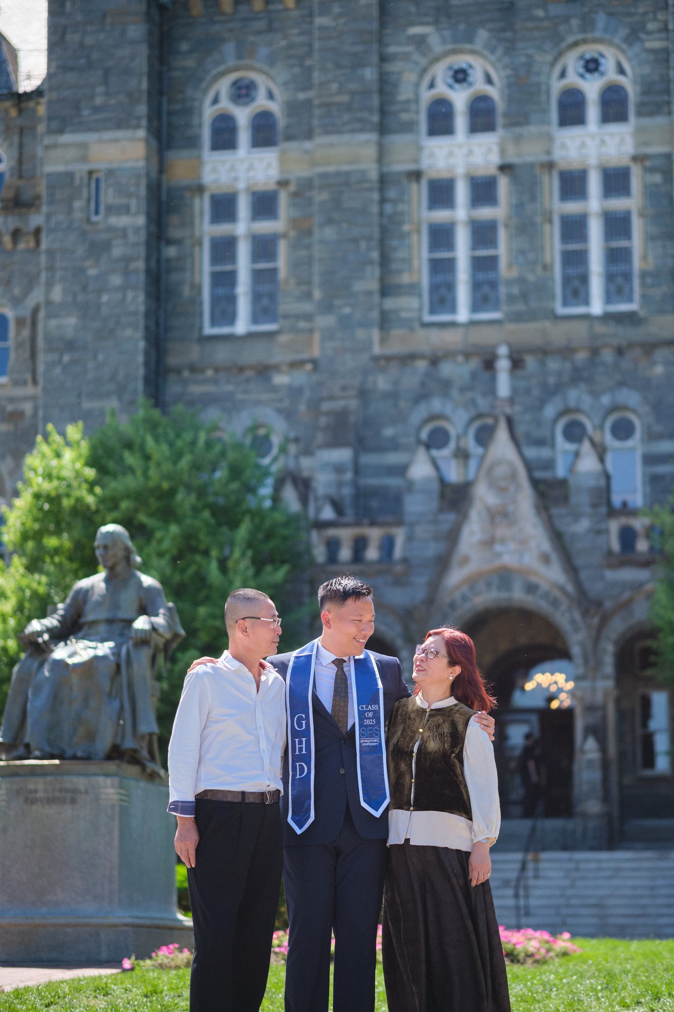 A graduation ceremony is taking place outdoors in front of a historic stone building with arched windows. Three people, one male graduate wearing a gown and sash, and two adults, are standing together and smiling, with the graduate in the middle. The