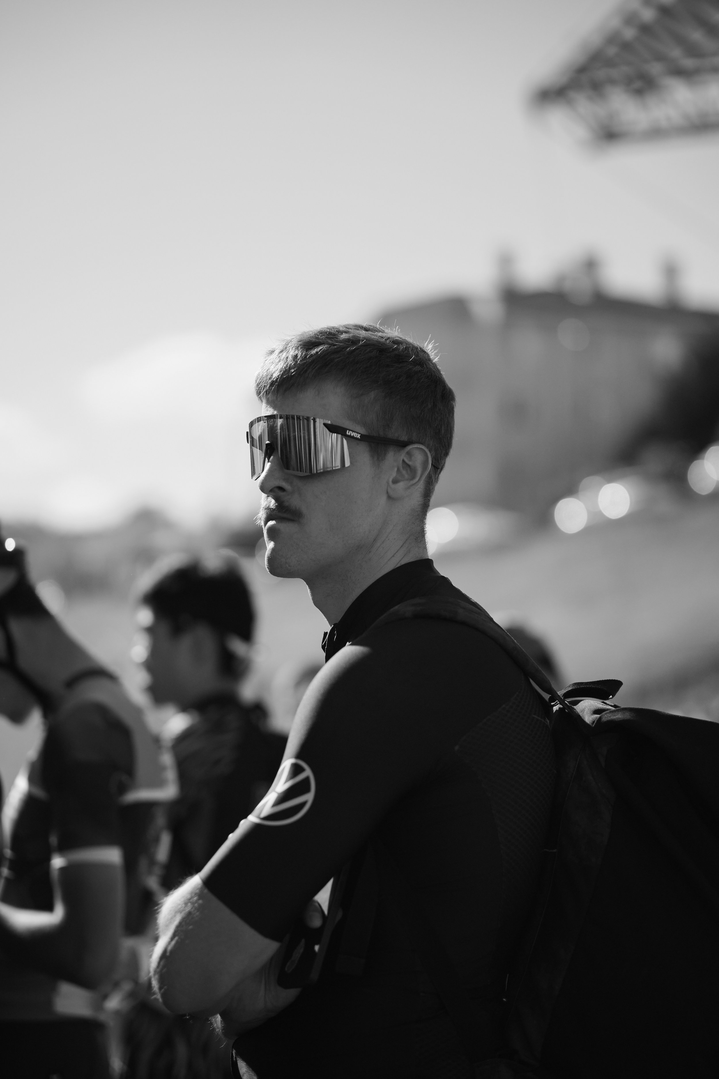 A young man with short hair wearing reflective sunglasses and a black sports shirt, standing outdoors among other cyclists, with a blurred building and structure in the background.