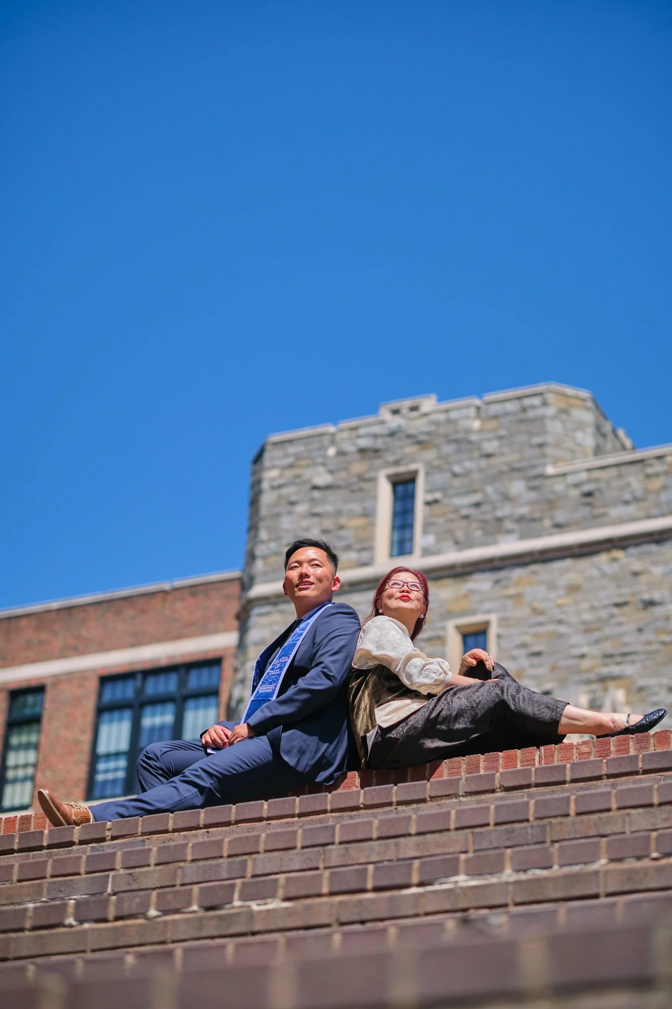 Two people sitting on brick steps in front of a stone building, with a bright blue sky overhead.