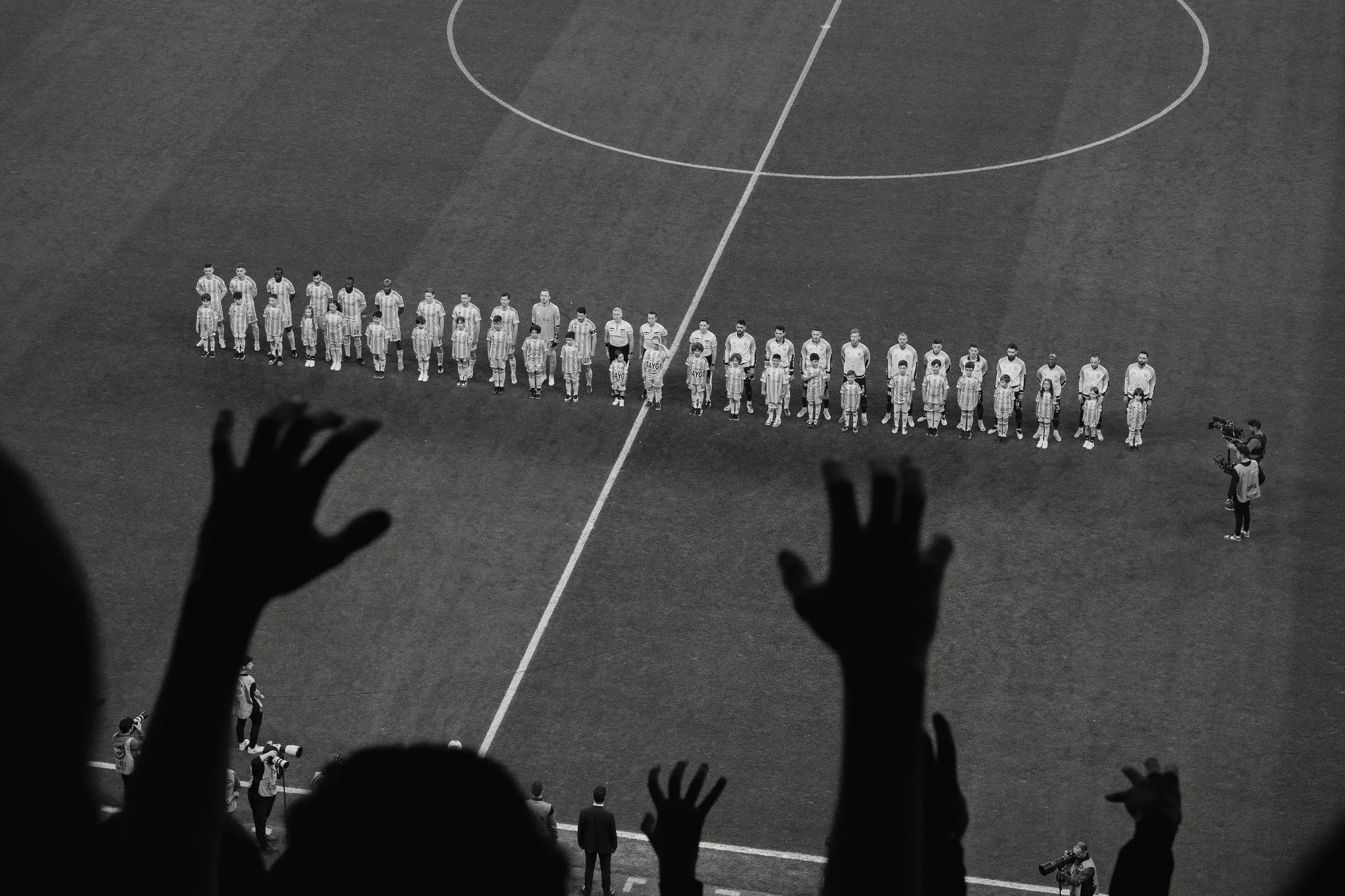 Black and white photo of a group of soccer players standing on a soccer field, viewed from the stands with hands and arms raised in the foreground.