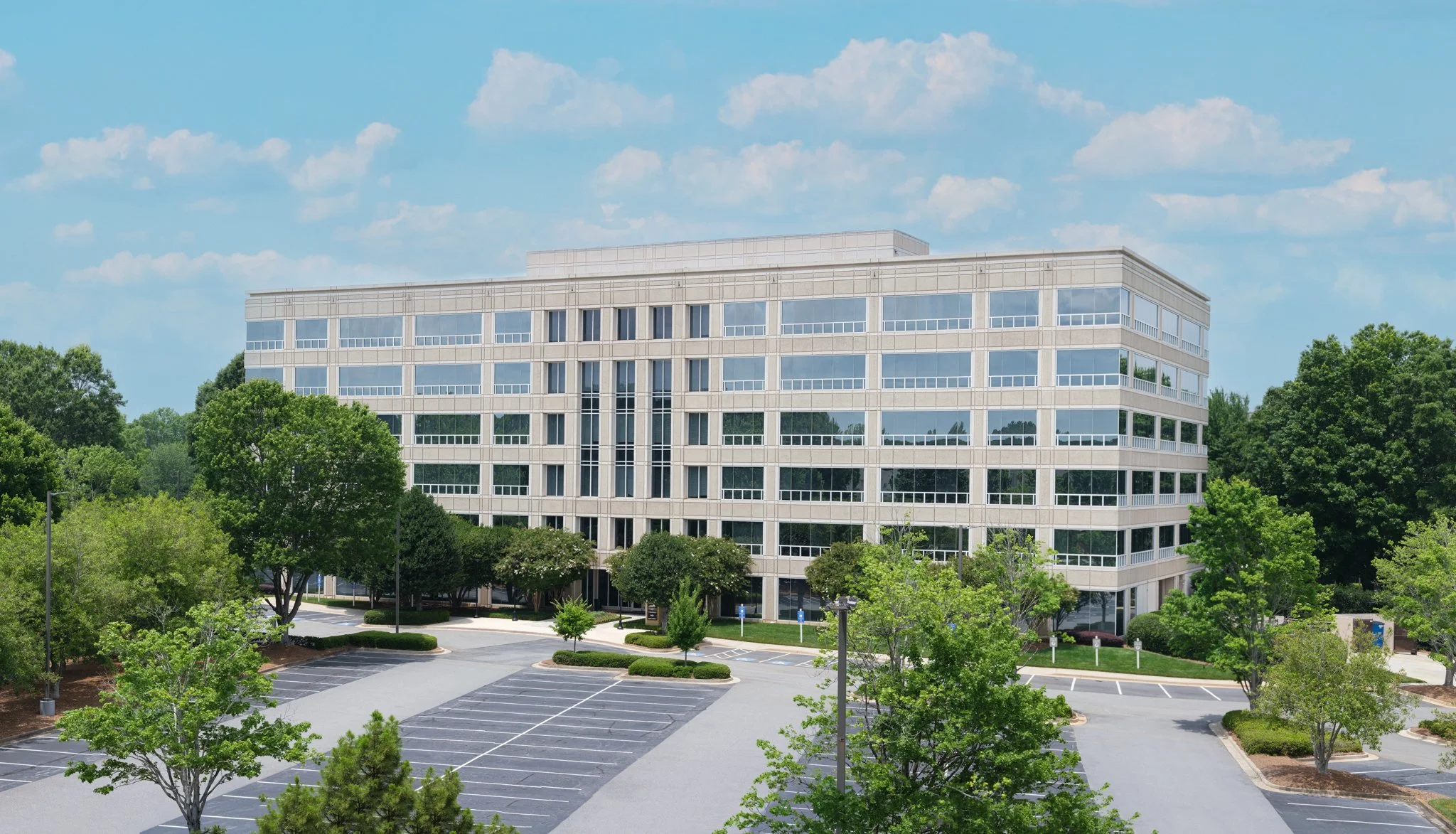 A modern office building with multiple floors and large glass windows, surrounded by green trees and an empty parking lot under a partly cloudy sky. | Real Estate Photography | Alpharetta | Milton | Buckhead | Atlanta | Drone | Northwinds | Architect