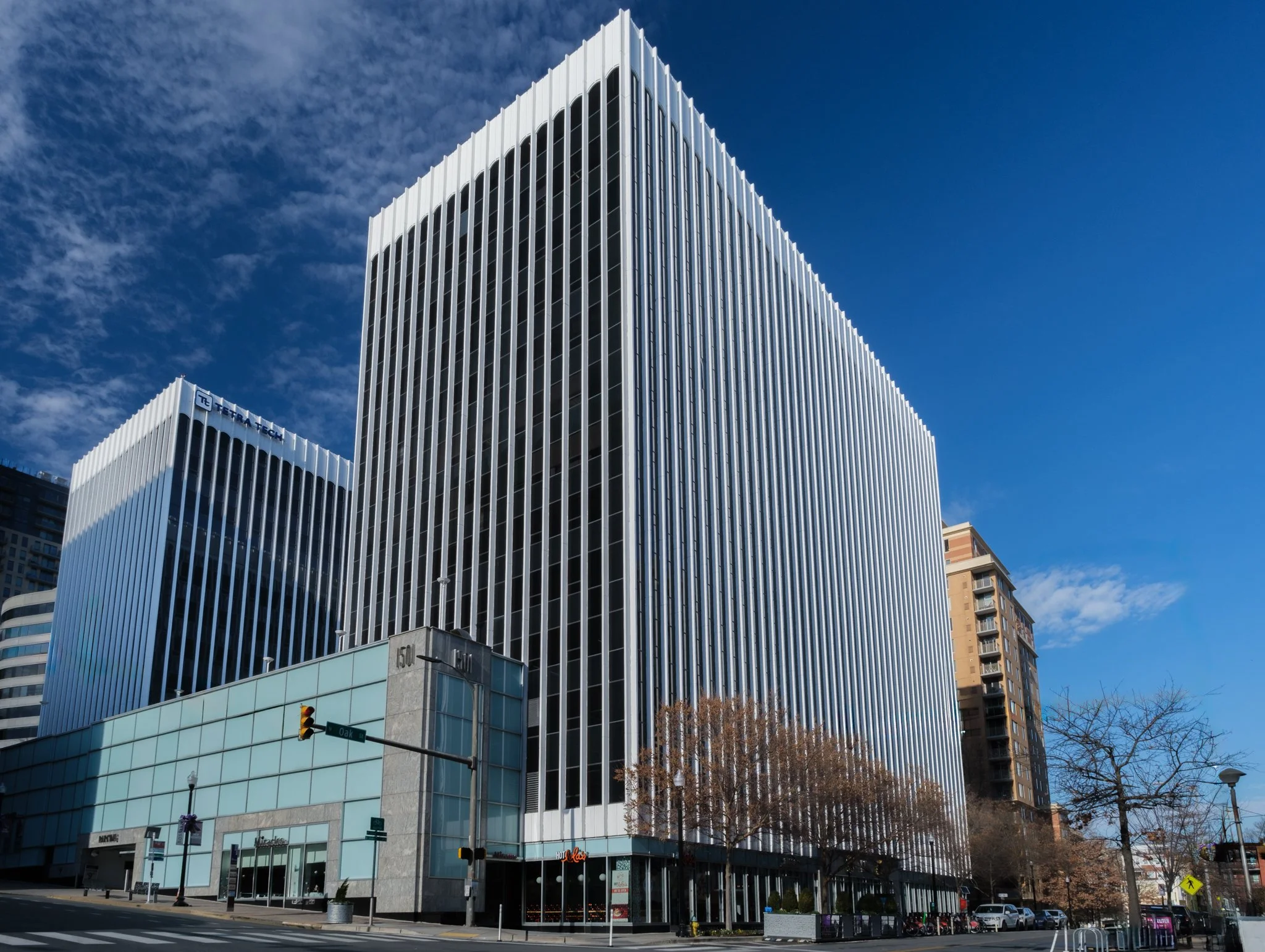 A modern high-rise office building with white vertical fins on its facade, standing on a city street with traffic lights and sidewalks, under a partly cloudy blue sky.