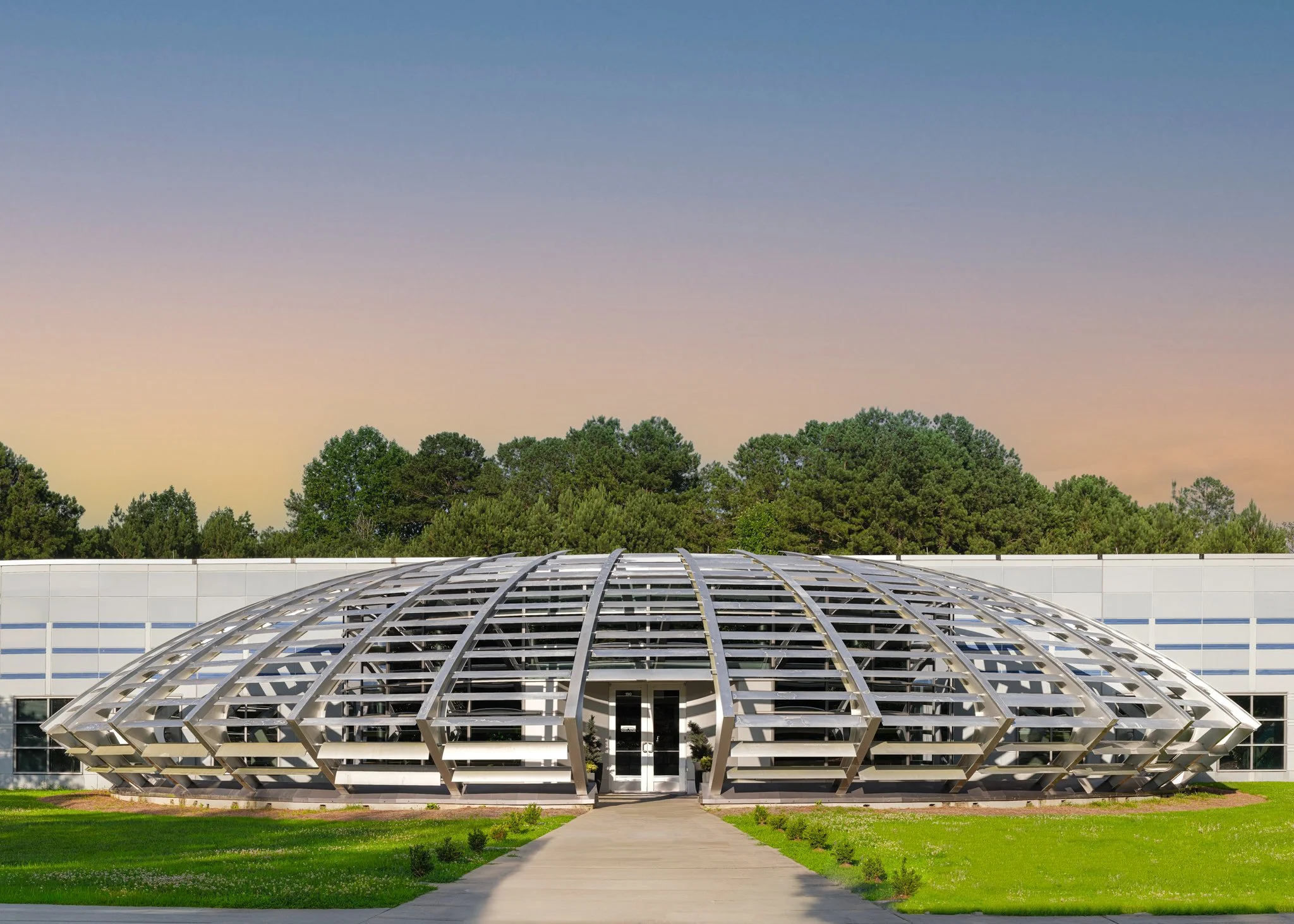 Modern building with a curved metal and glass roof in front of a green lawn and trees, under a clear sky at sunset. | Real Estate Photography | Alpharetta | Milton | Buckhead | Atlanta | Drone | Video | Media | Luxury | Digital Ignition Building | Ar
