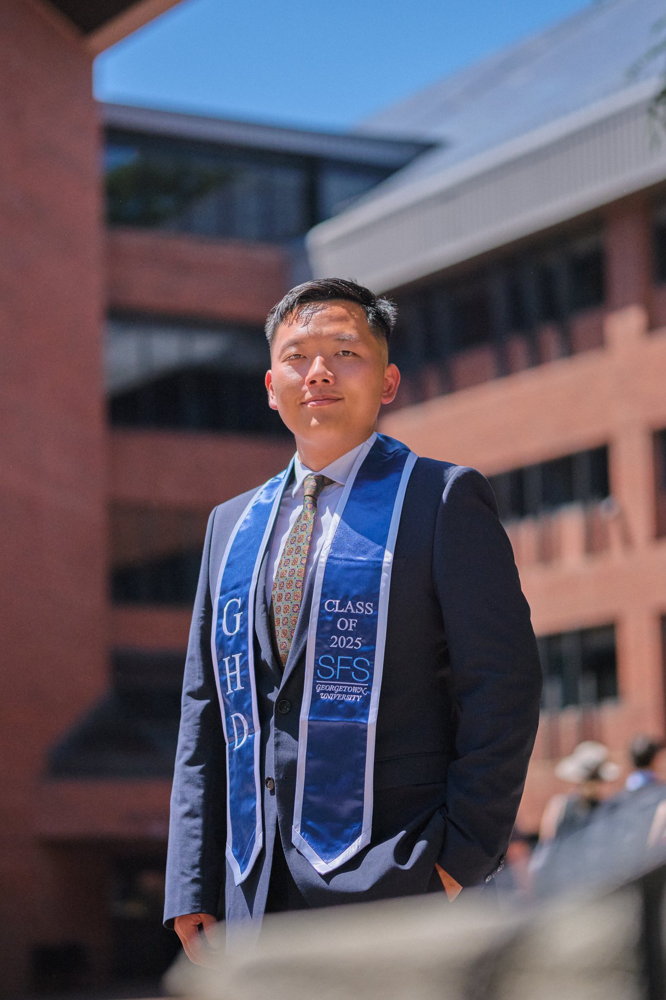 Young man in formal suit wearing honor cords and a stole that says 'Class of 2025' from George Washington University, outdoors during the evening.