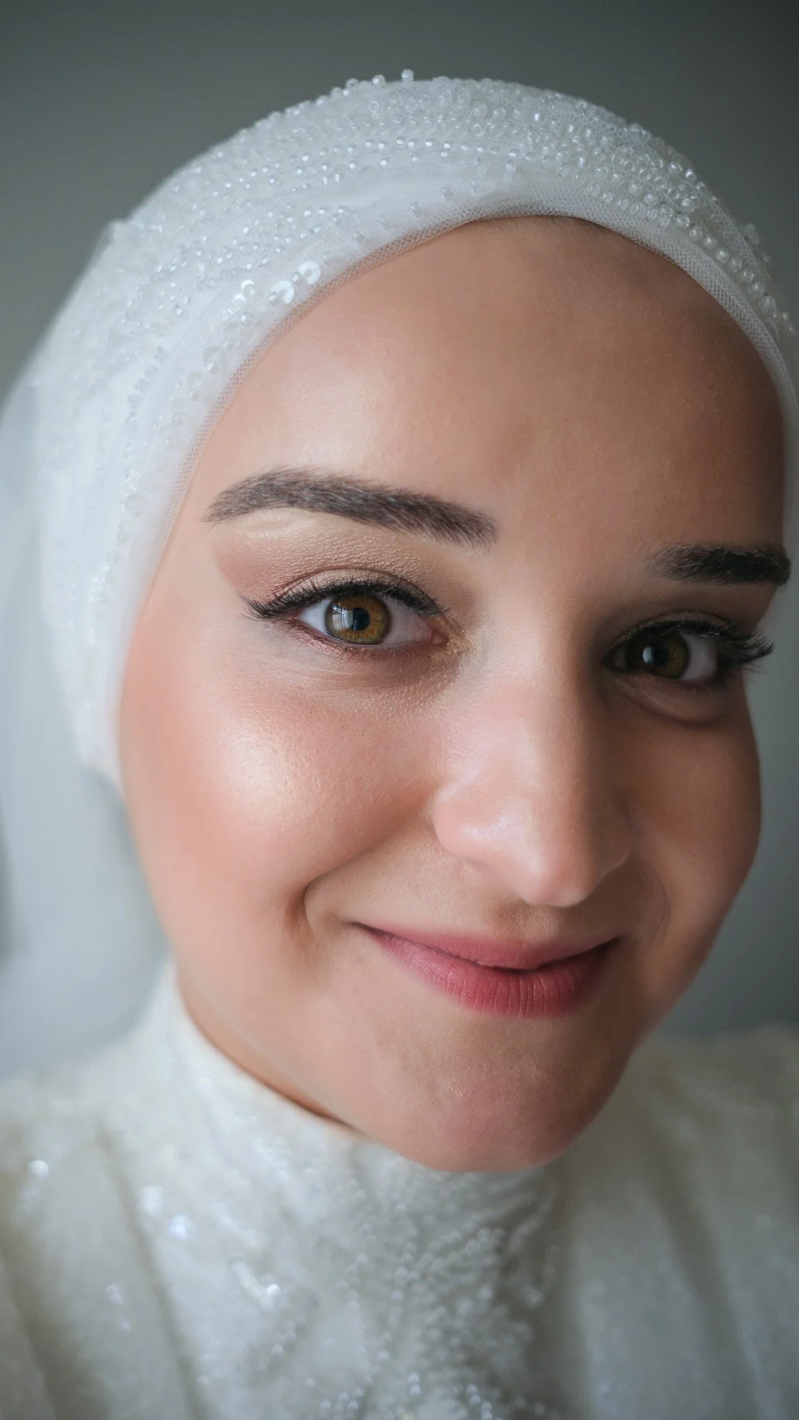 Close-up of a woman wearing a white headscarf decorated with small pearls, showing her face with makeup and a slight smile.