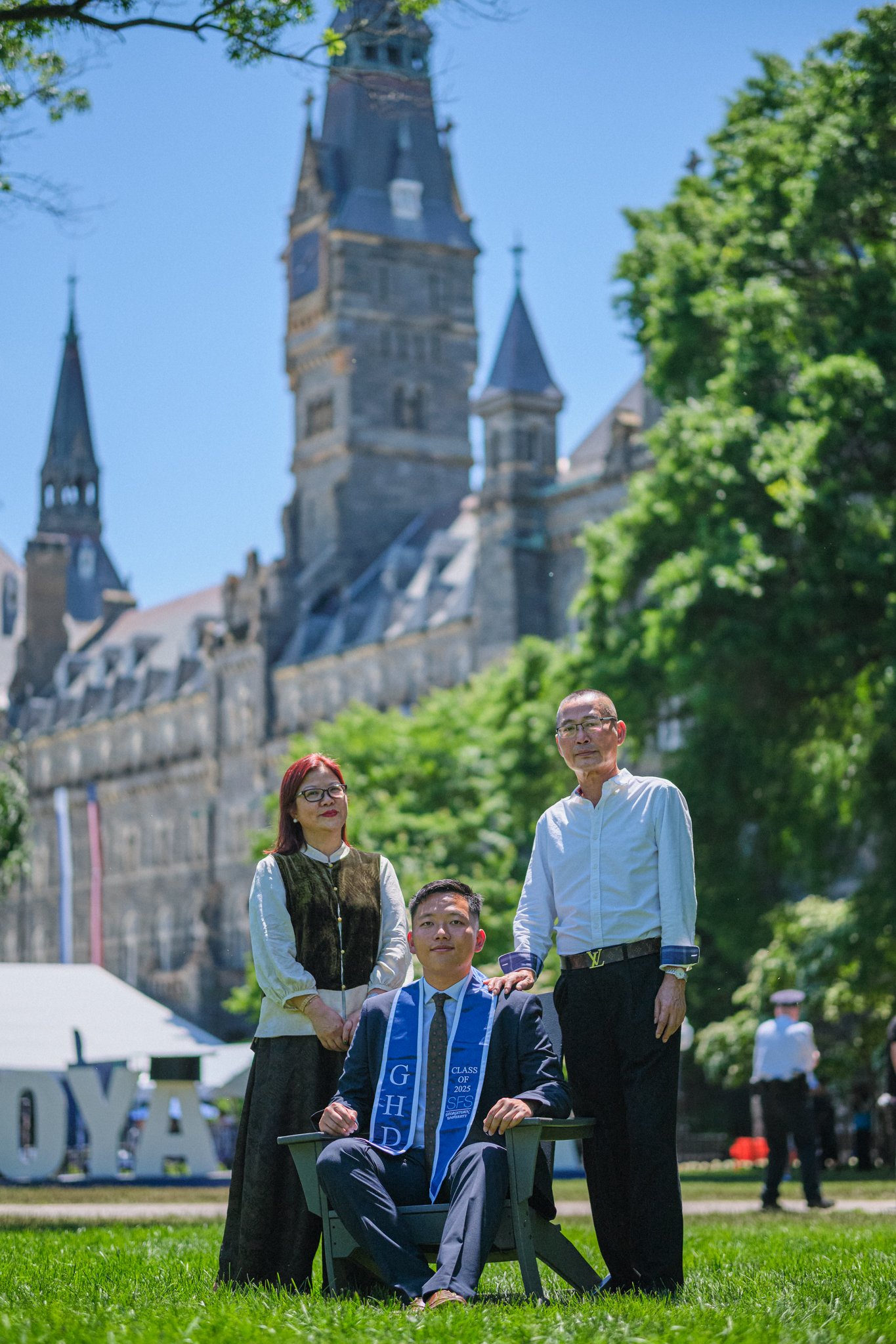 Four people at a graduation ceremony outdoors with a historic castle in the background. One person is seated in a chair wearing a graduation stole, and three people are standing behind him, two men and a woman, all dressed in formal attire.