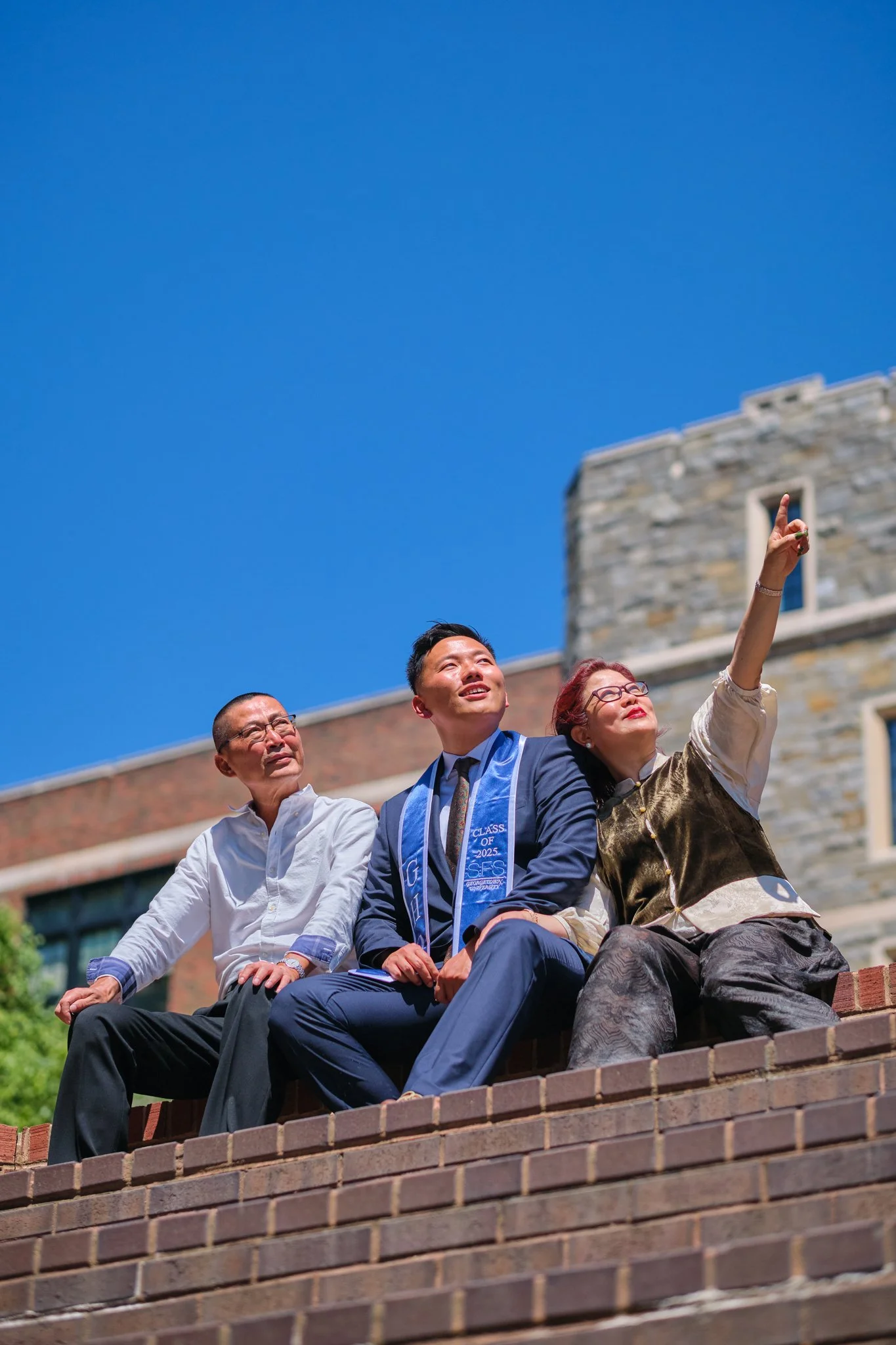 Four people sitting on a brick ledge outdoors, with a building and a clear blue sky in the background. One woman is pointing upward, and another person is wearing a graduation sash.