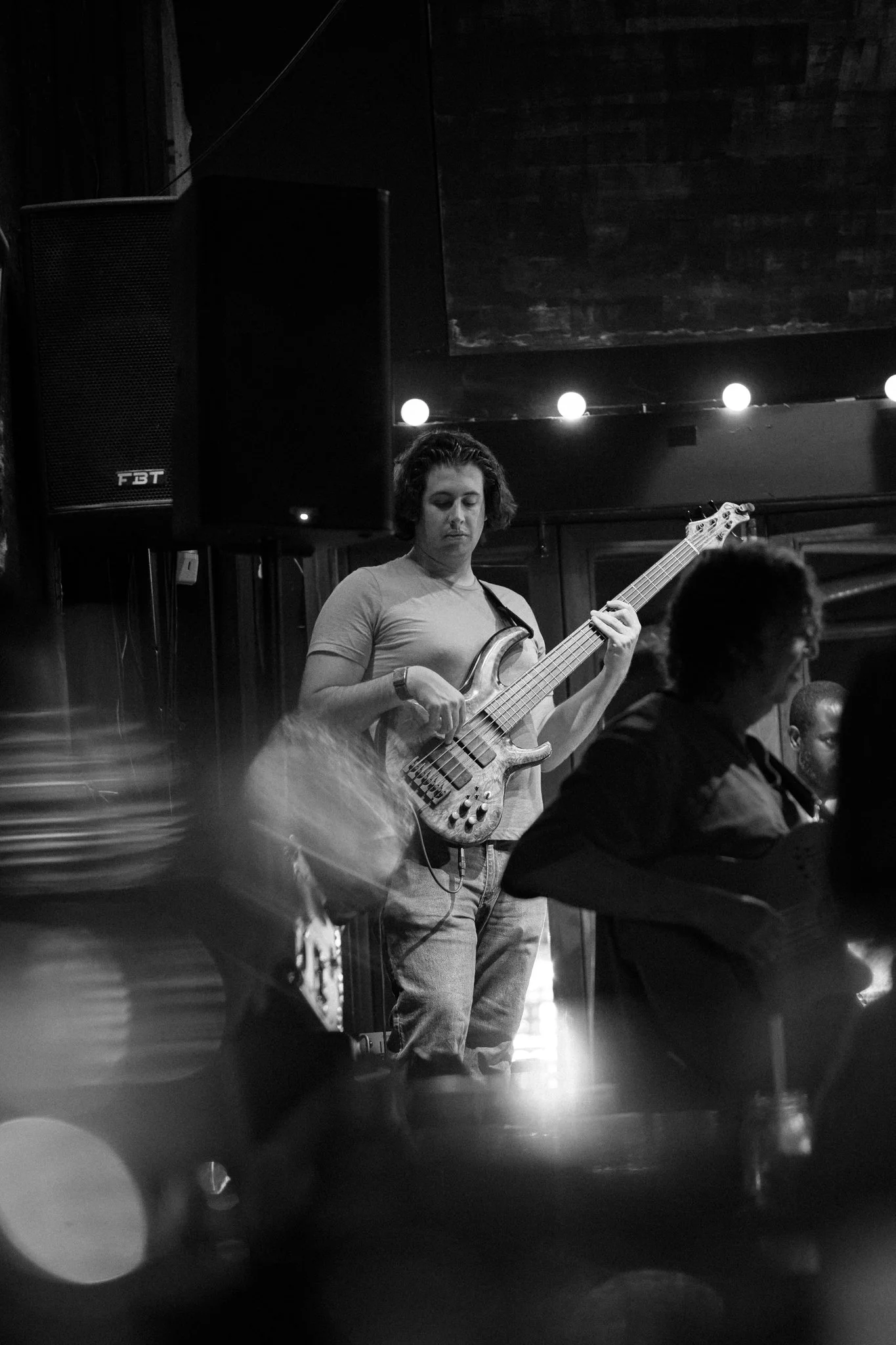 A woman with short curly hair playing a bass guitar on stage in a dimly lit venue, surrounded by audience members, with a large speaker behind her and string lights overhead.