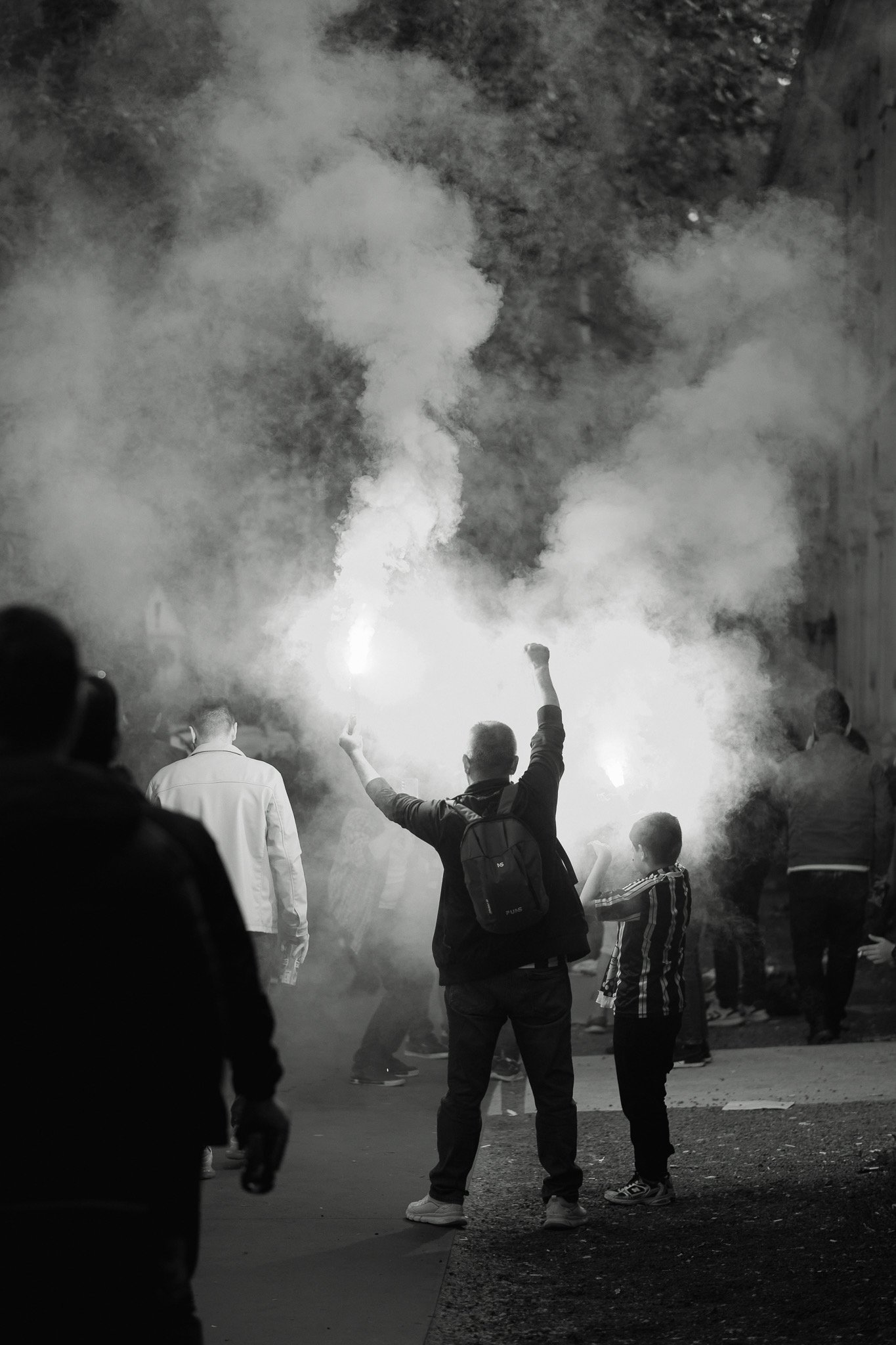 People gathered outdoors at night, some holding flares or smoke bombs, with visible smoke and bright light in the background.
