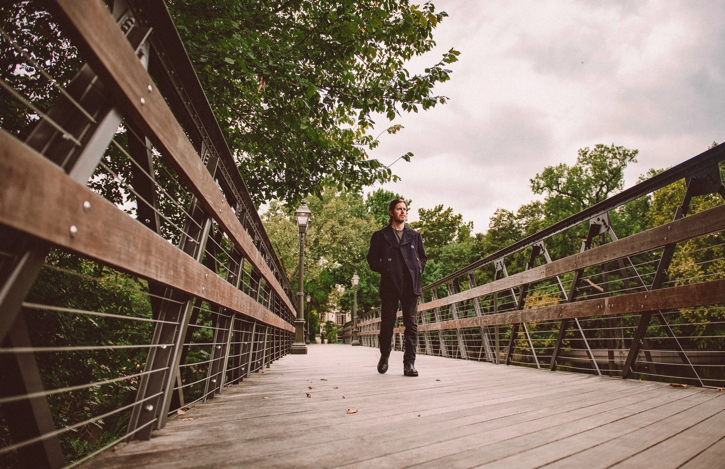 A man walking on a wooden bridge with metal railings in a park, surrounded by trees and overcast sky.