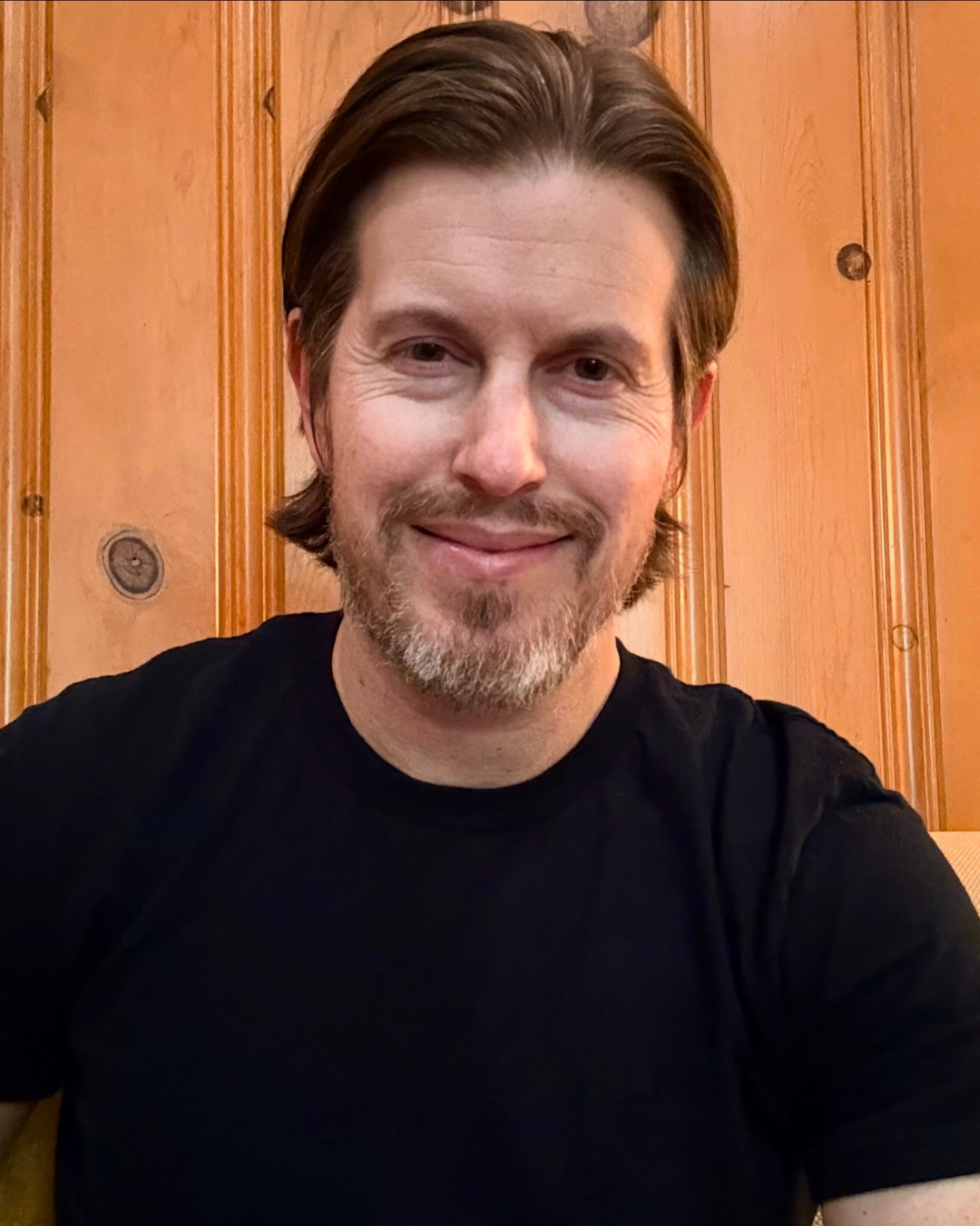 A middle-aged man with brown hair, a beard, and mustache smiling at the camera, wearing a black shirt, sitting in front of a wooden-paneled wall.