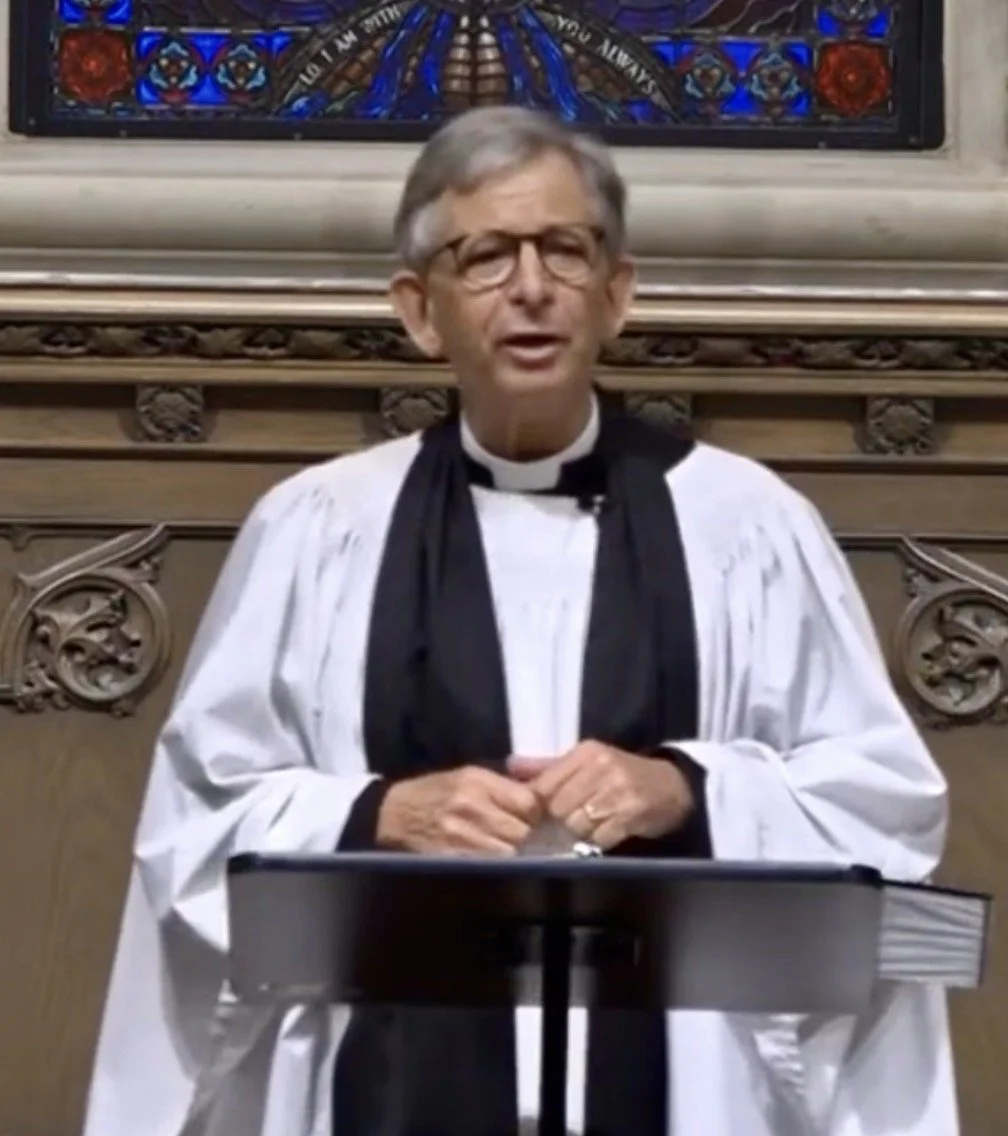 A priest wearing glasses and a traditional white clergy robe with a black stole, standing behind a lectern inside a church. A stained glass window is visible in the background.