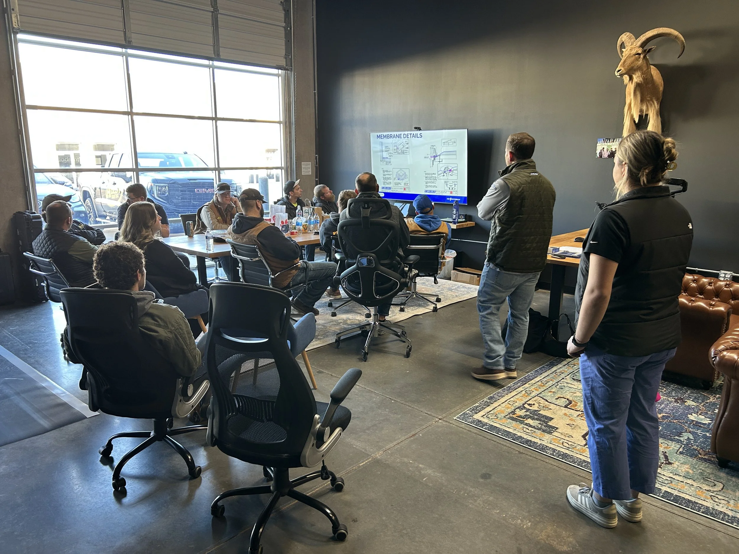 A group of people attending a presentation or seminar in an industrial-style room with large windows. The presenter is standing near a large screen displaying detailed diagrams, and there is a mounted animal skull or sculpture on the wall.