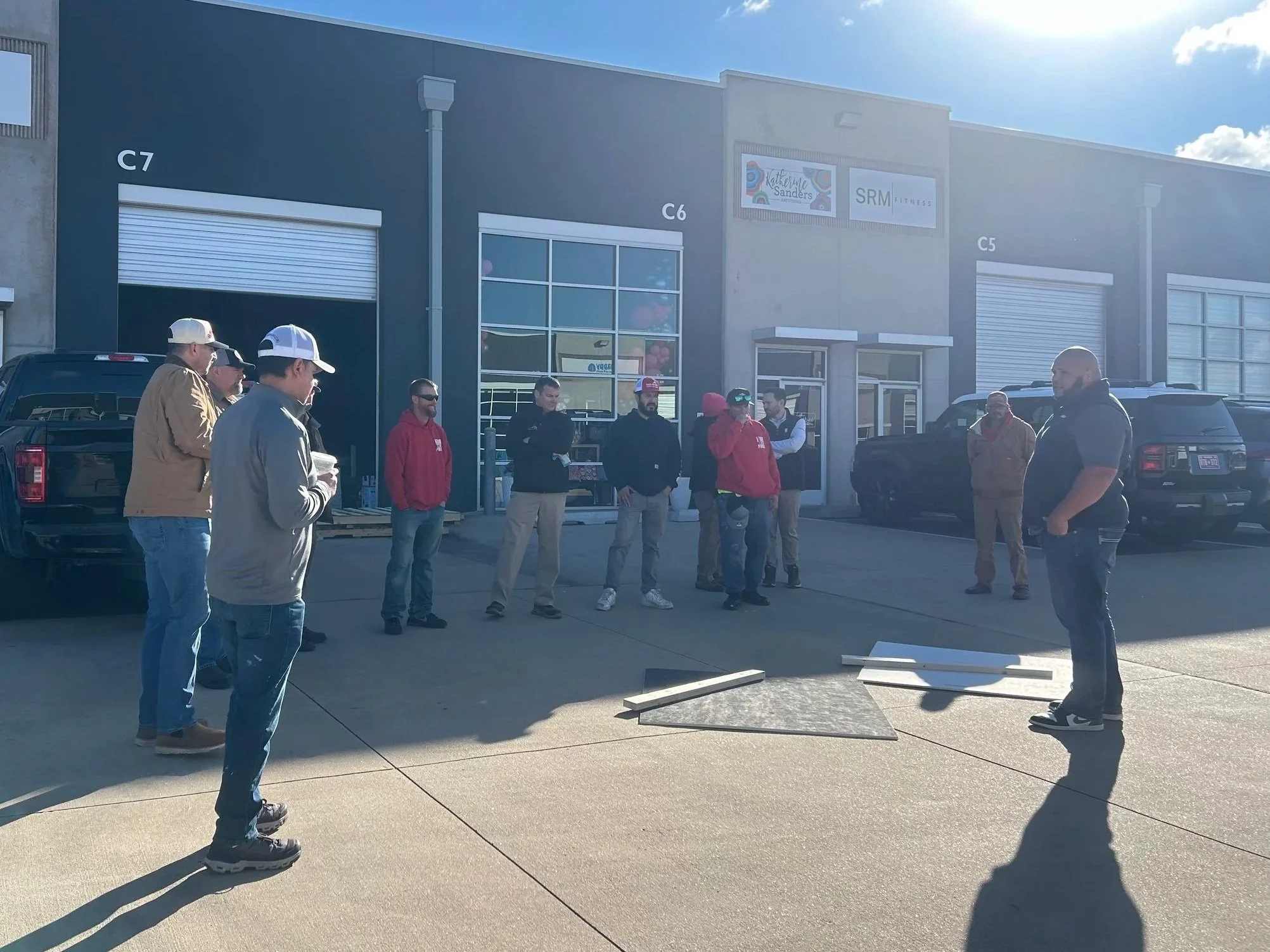 Group of people standing outside a commercial building with office or retail units, some wearing jackets and hats, near parked cars, with bright sunlight and a clear sky.
