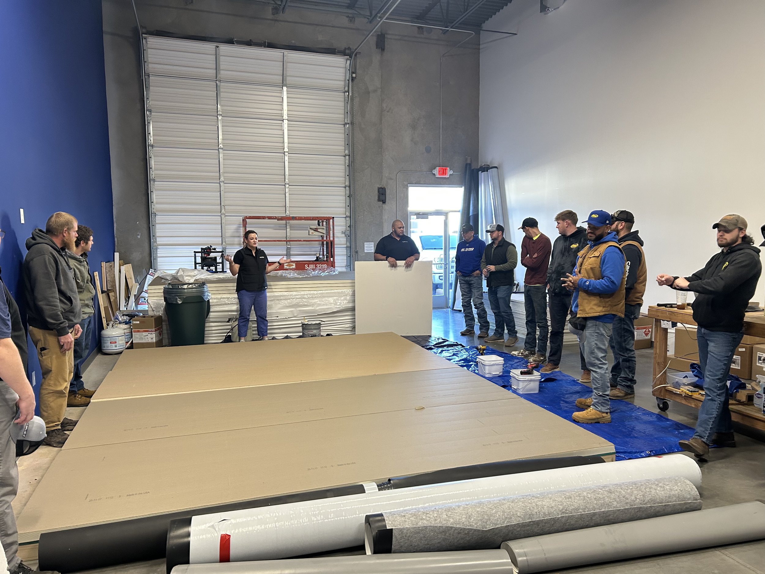 A group of people standing inside a warehouse or workshop, engaged in a discussion or presentation. There are large rolls of material, possibly vinyl or fabric, on a table and the floor. The space has a high ceiling with a large garage door, and an instructor is speaking to the group.