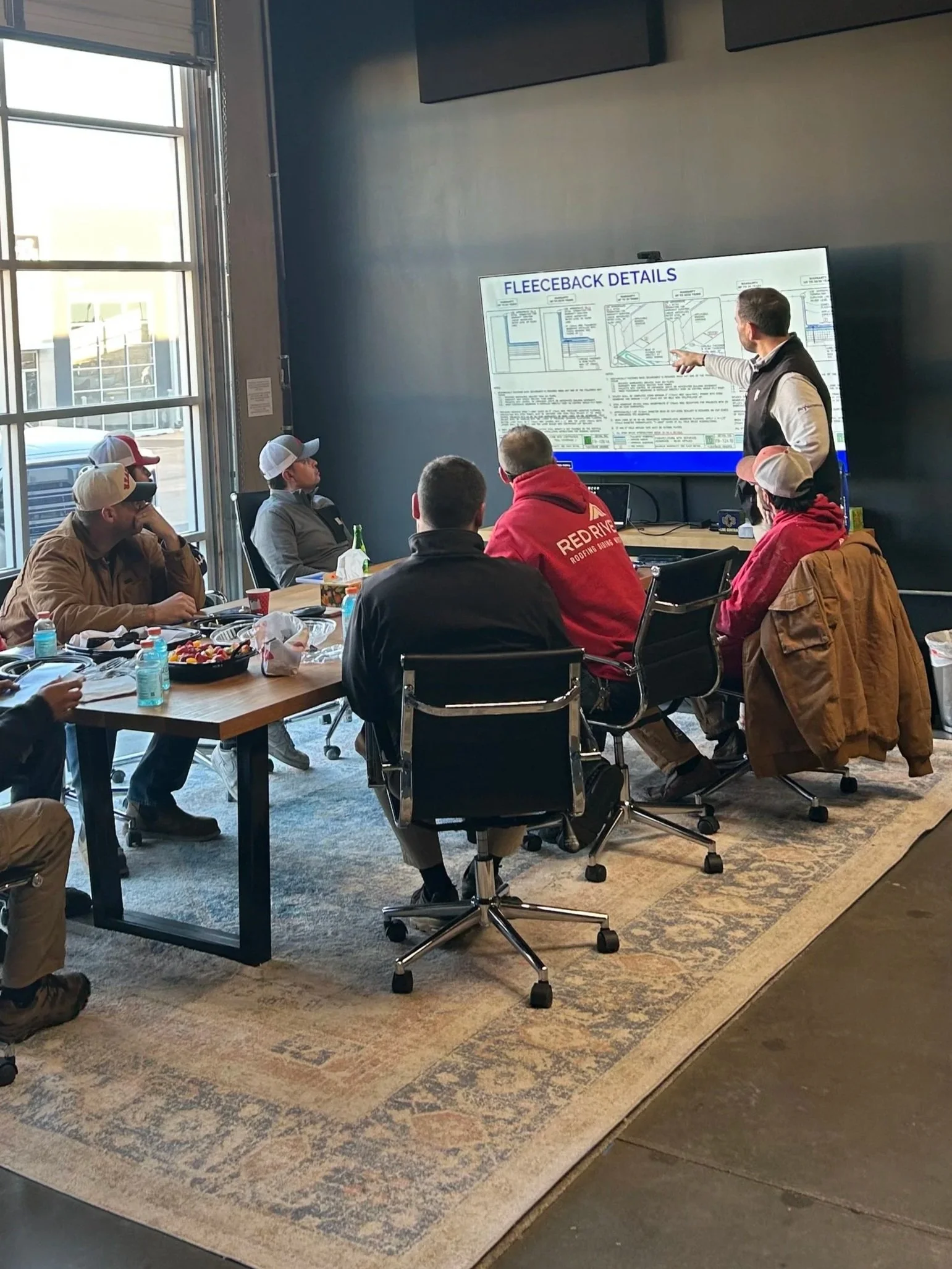 A man in a black vest is giving a presentation to a group of people seated at a long table. The audience includes both men and women, some wearing hats and jackets. The room has large windows allowing natural light, and there is a large display screen showing a detailed chart titled "FLECEBACK DETAILS". The table is filled with snacks, drinks, and personal items.