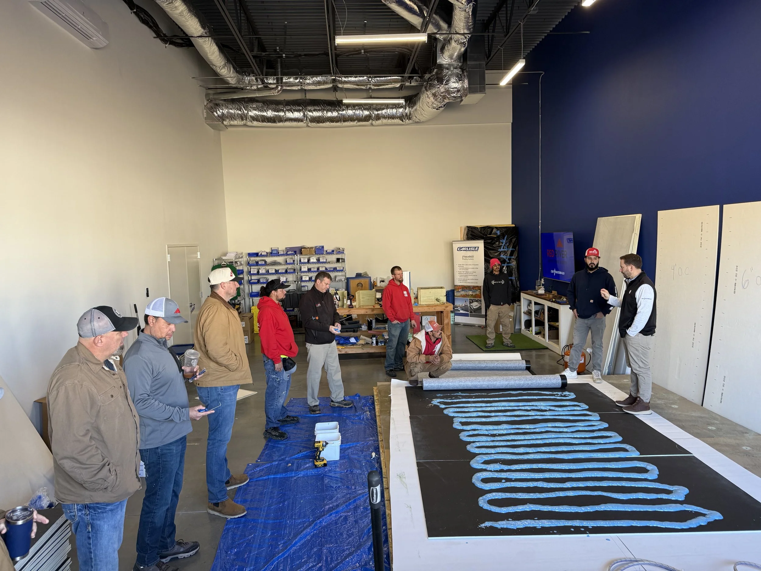 A group of people gathered inside a large industrial-style room, watching a demonstration involving large sheets of material on the floor, with some blue and black painted areas, and long blue tubes or hoses laid out in parallel rows.