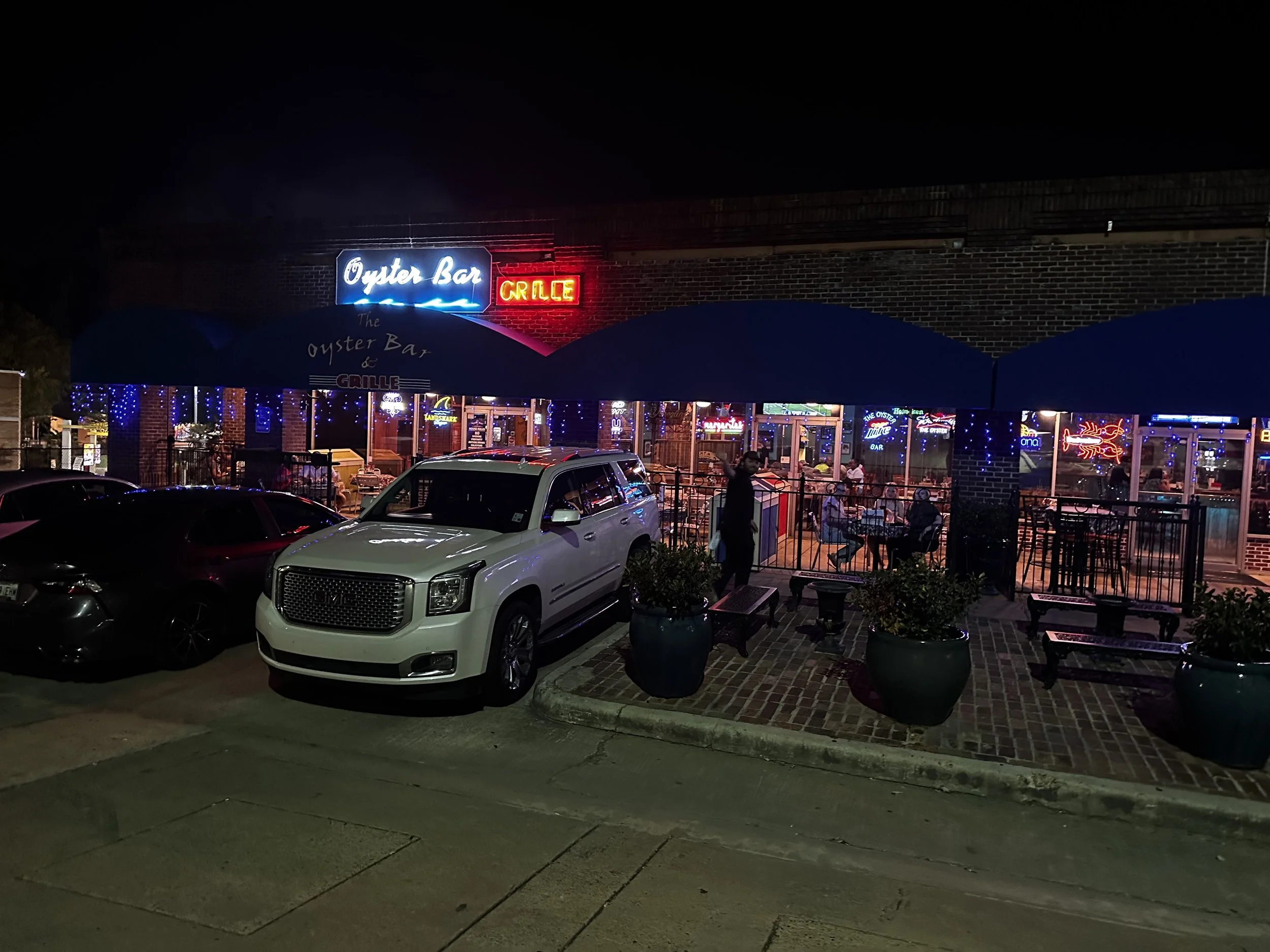 Night view of a restaurant called The Oyster Bar & Grille with neon signs, outdoor seating, and parked cars in front.