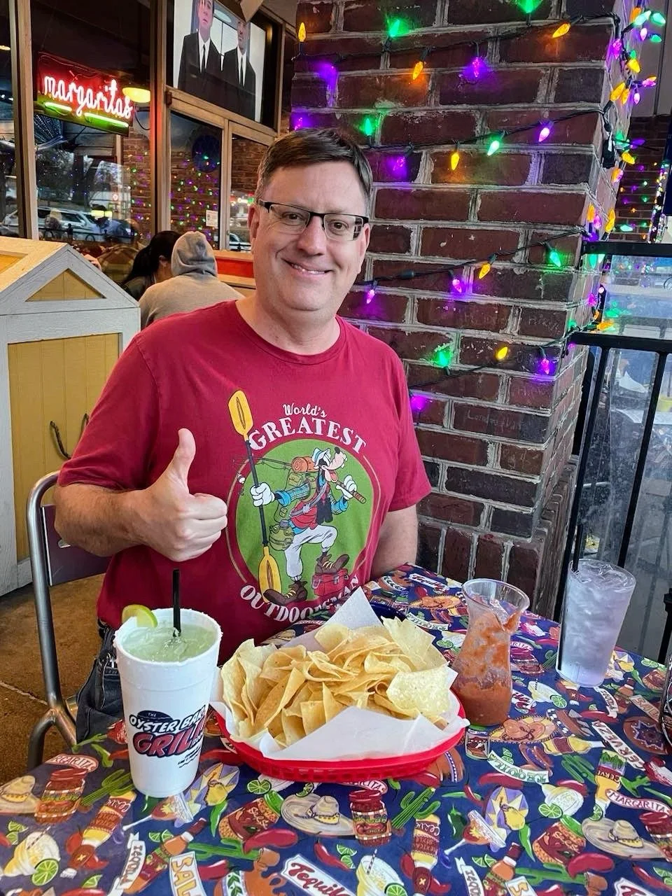 Man sitting at a decorated restaurant table with chips and drinks, giving a thumbs up and smiling, wearing glasses and a red T-shirt with an outdoor adventure design. The table is covered with a colorful taco-themed tablecloth, and the background fea