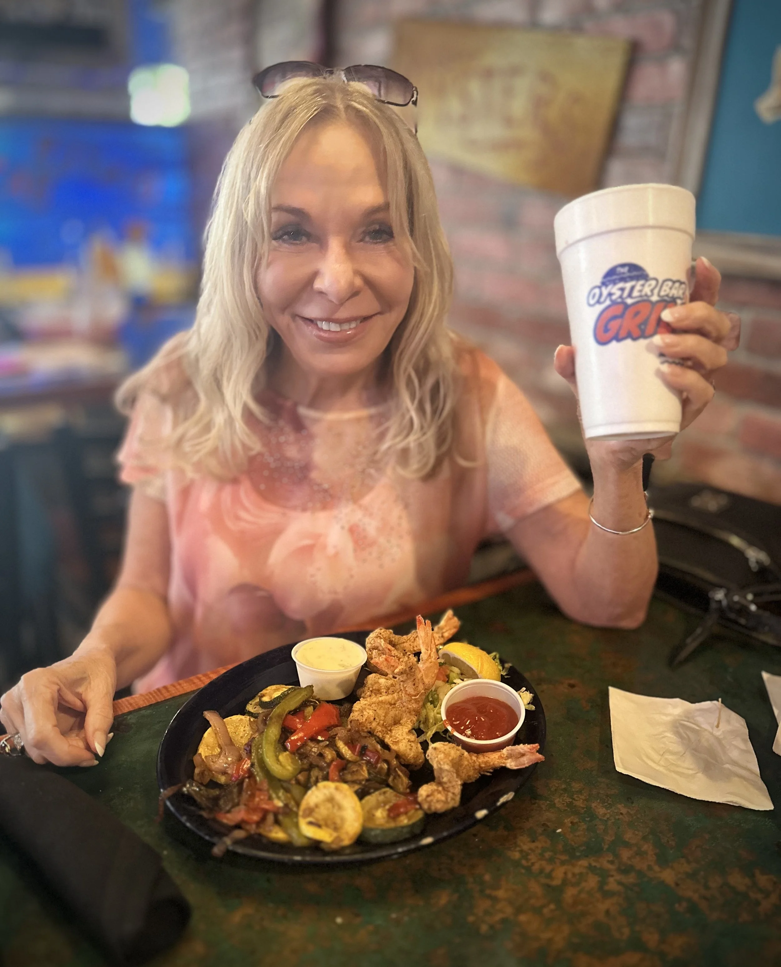 A woman with blonde hair and sunglasses on her head smiling and holding a white cup with the logo 'Oyster Bar Grill' in a restaurant. In front of her, there is a sizzling platter of fried seafood, vegetables, and lemon slices, with small containers o