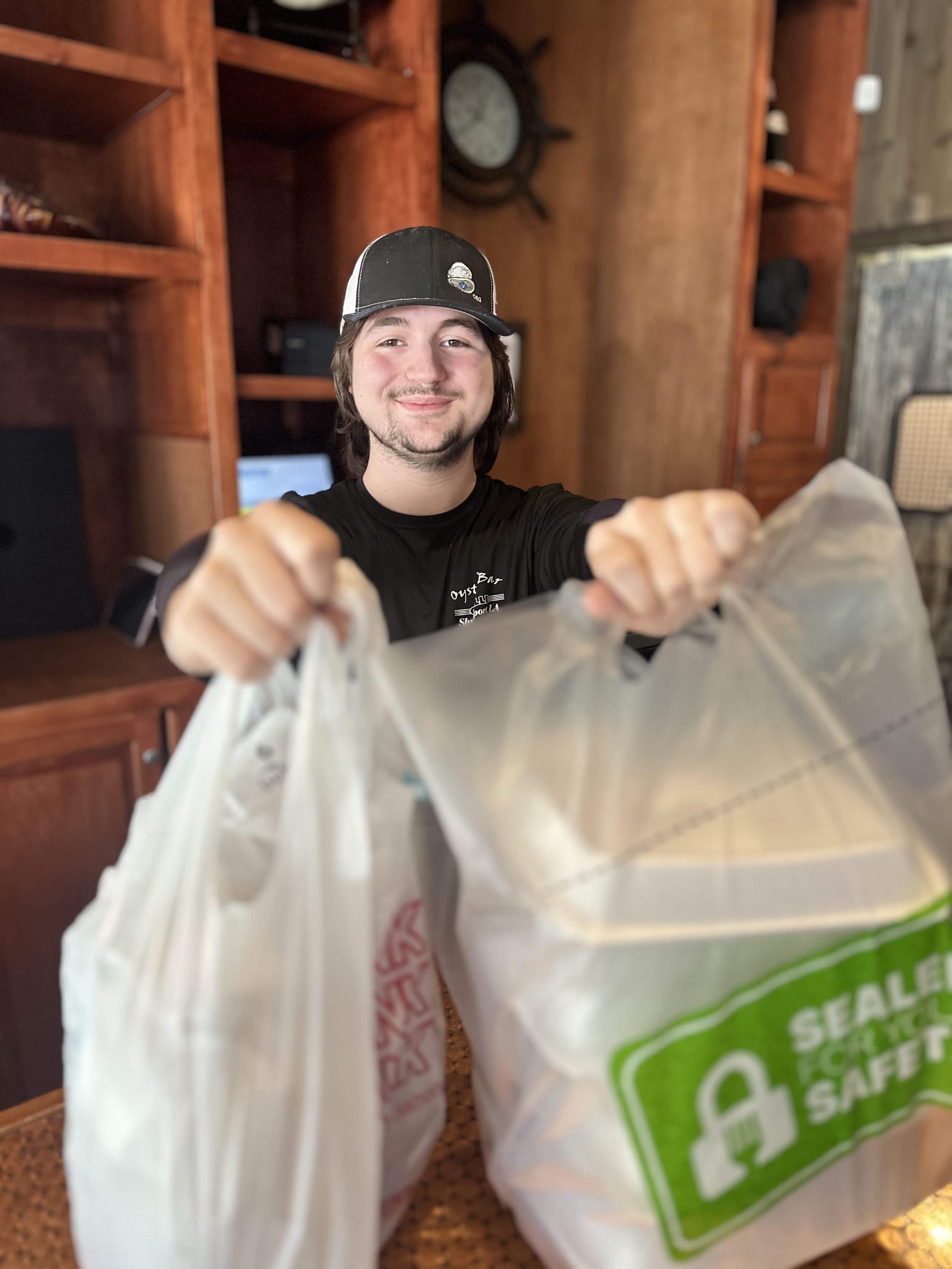 A man wearing a baseball cap and black shirt smiling at the camera while holding a white shopping bag in front of a wooden backdrop with shelves and a wall clock.