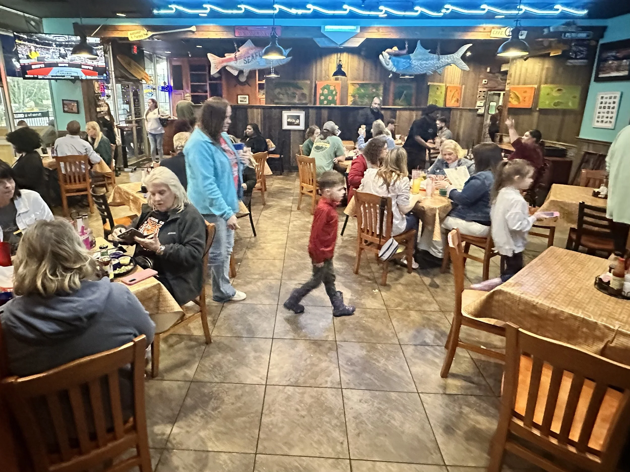The image shows a busy seafood restaurant filled with many people dining and chatting at wooden tables. There are decorations of large fish mounted on the walls, a television mounted on the wall, and a staff member walking through the restaurant. The