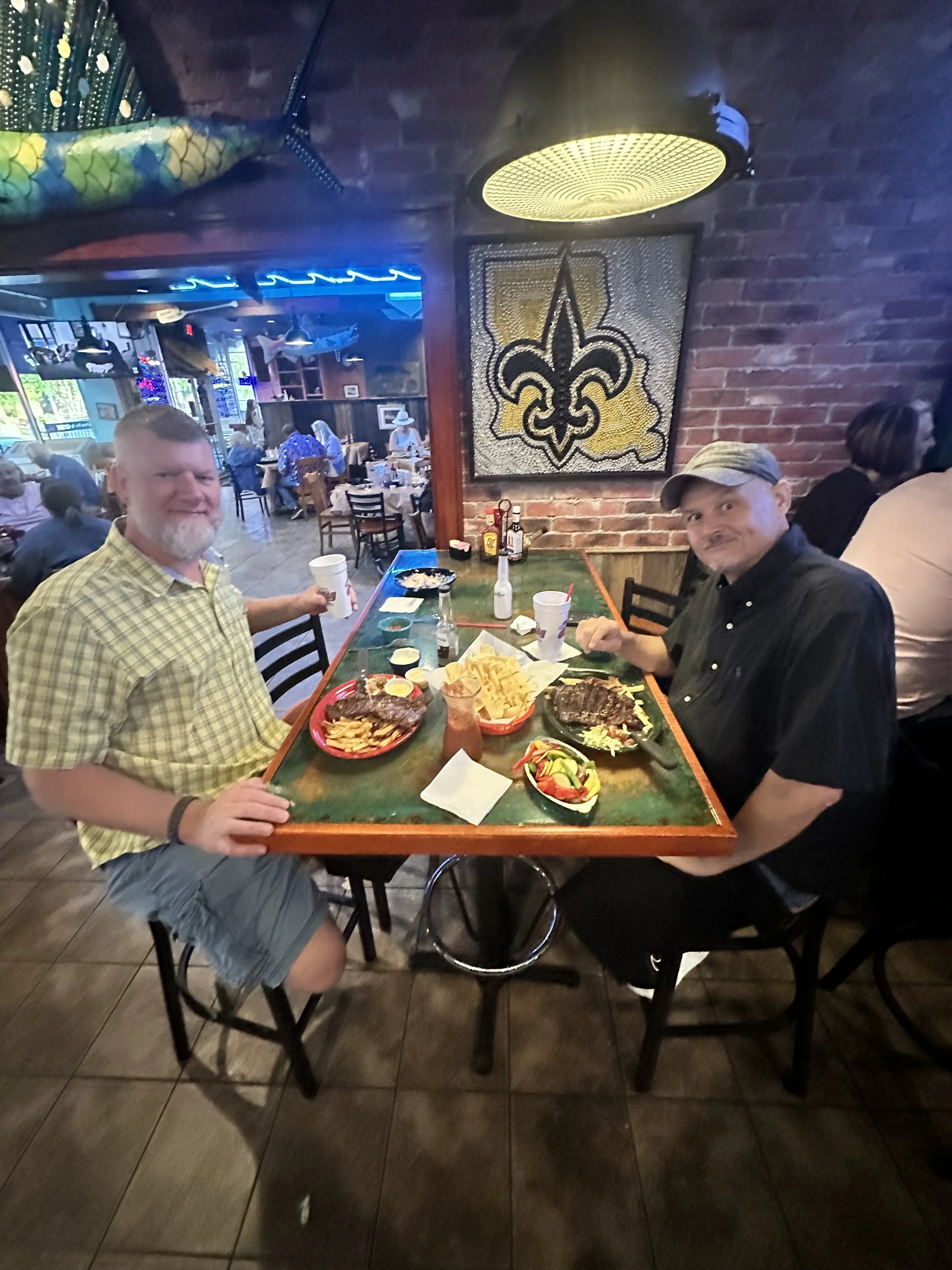 Two men sitting at a restaurant table with food and drinks, in a room with brick walls, a New Orleans Saints logo art piece, and several other customers visible in the background.