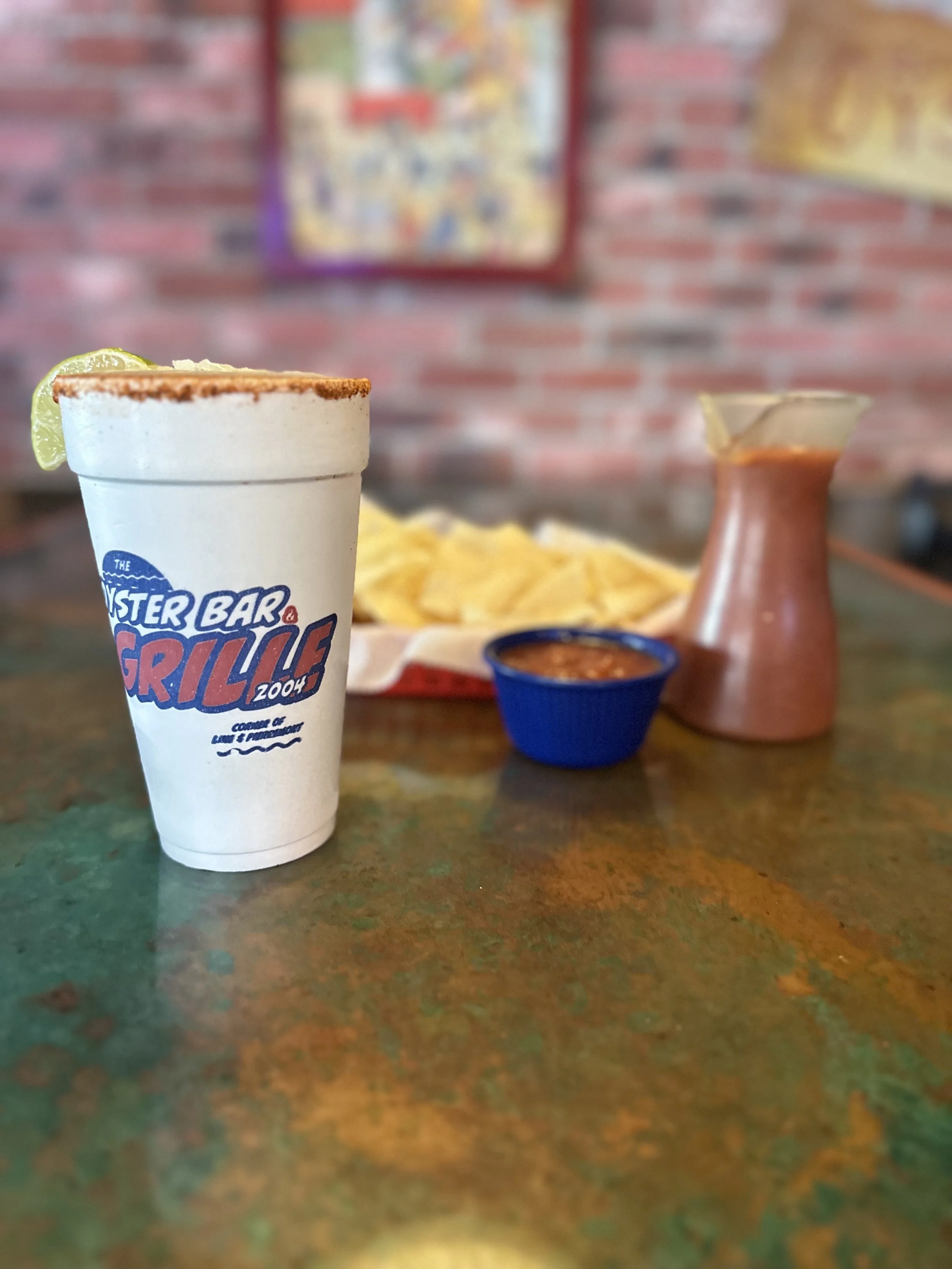 A Mexican meal with nachos, salsa, a small pitcher of sauce, and a salt shaker on a table in a restaurant with brick walls and colorful posters in the background.
