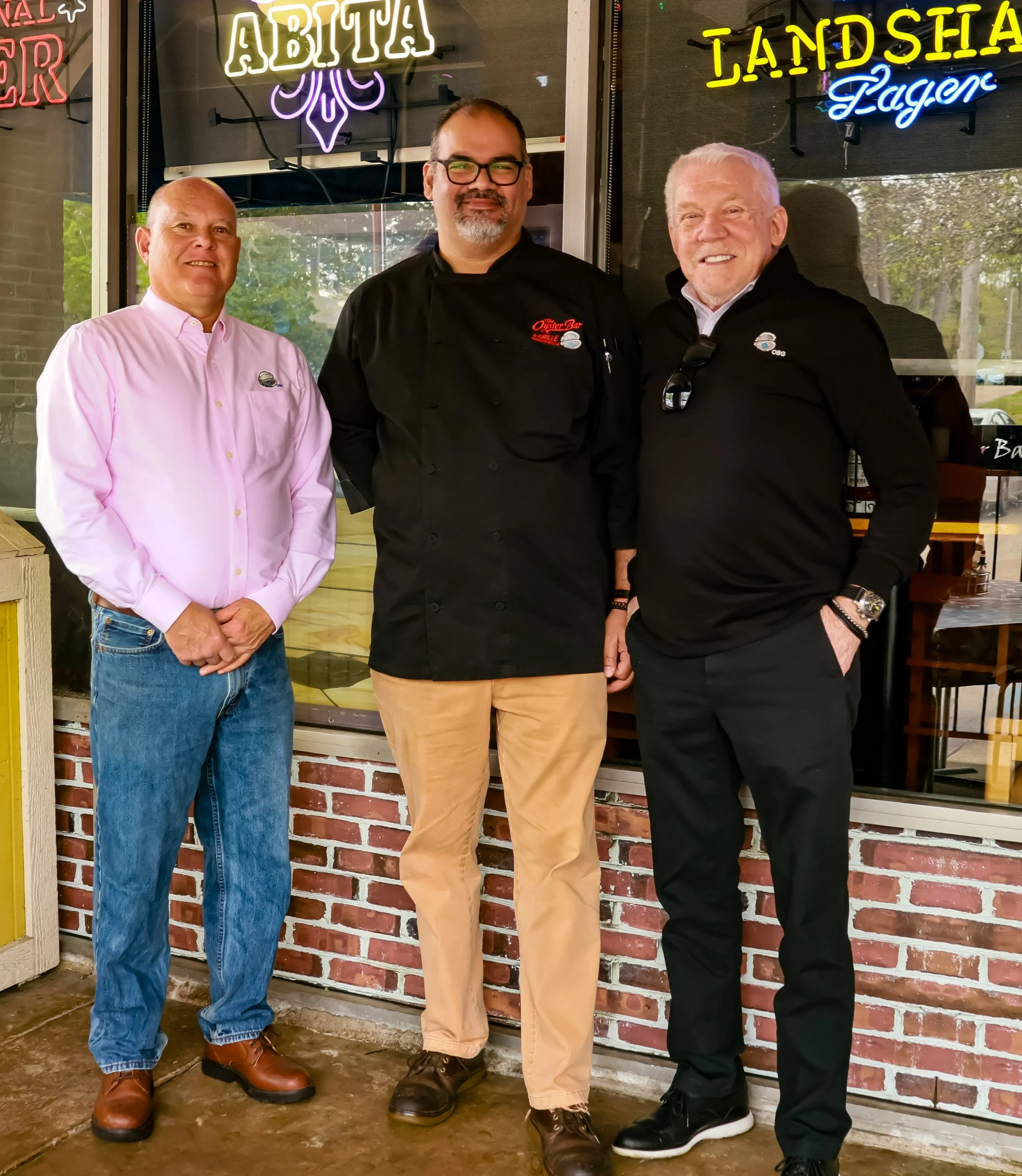 Three men standing together in front of a restaurant, two wearing black and one wearing pink, with neon signs above them reading 'Abita' and 'Landshark Lager'.