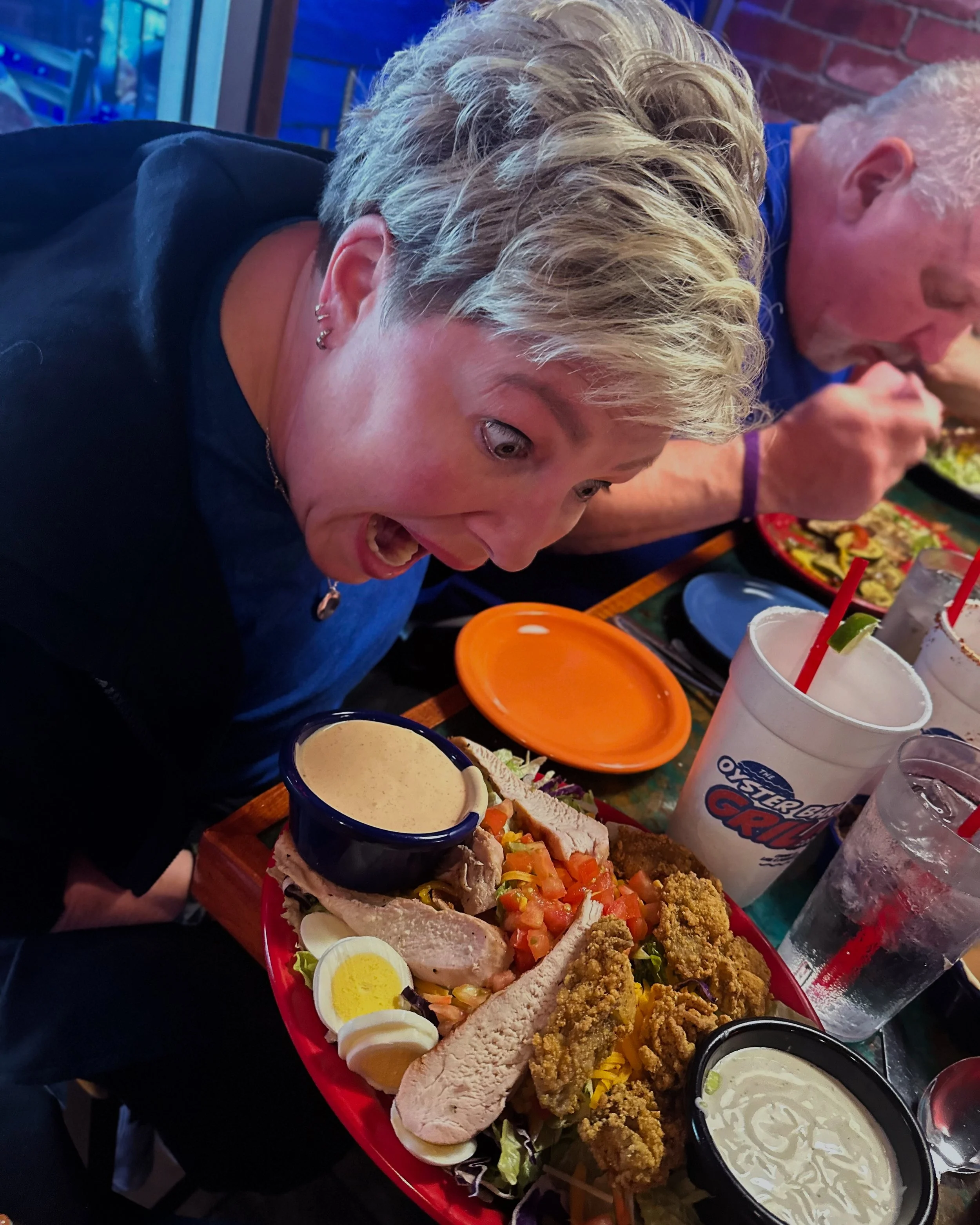 A person with short, light-colored hair enjoying a meal at a restaurant, with a plate full of various foods including fried chicken, sliced vegetables, boiled eggs, and bread, alongside drinks and condiments on the table.