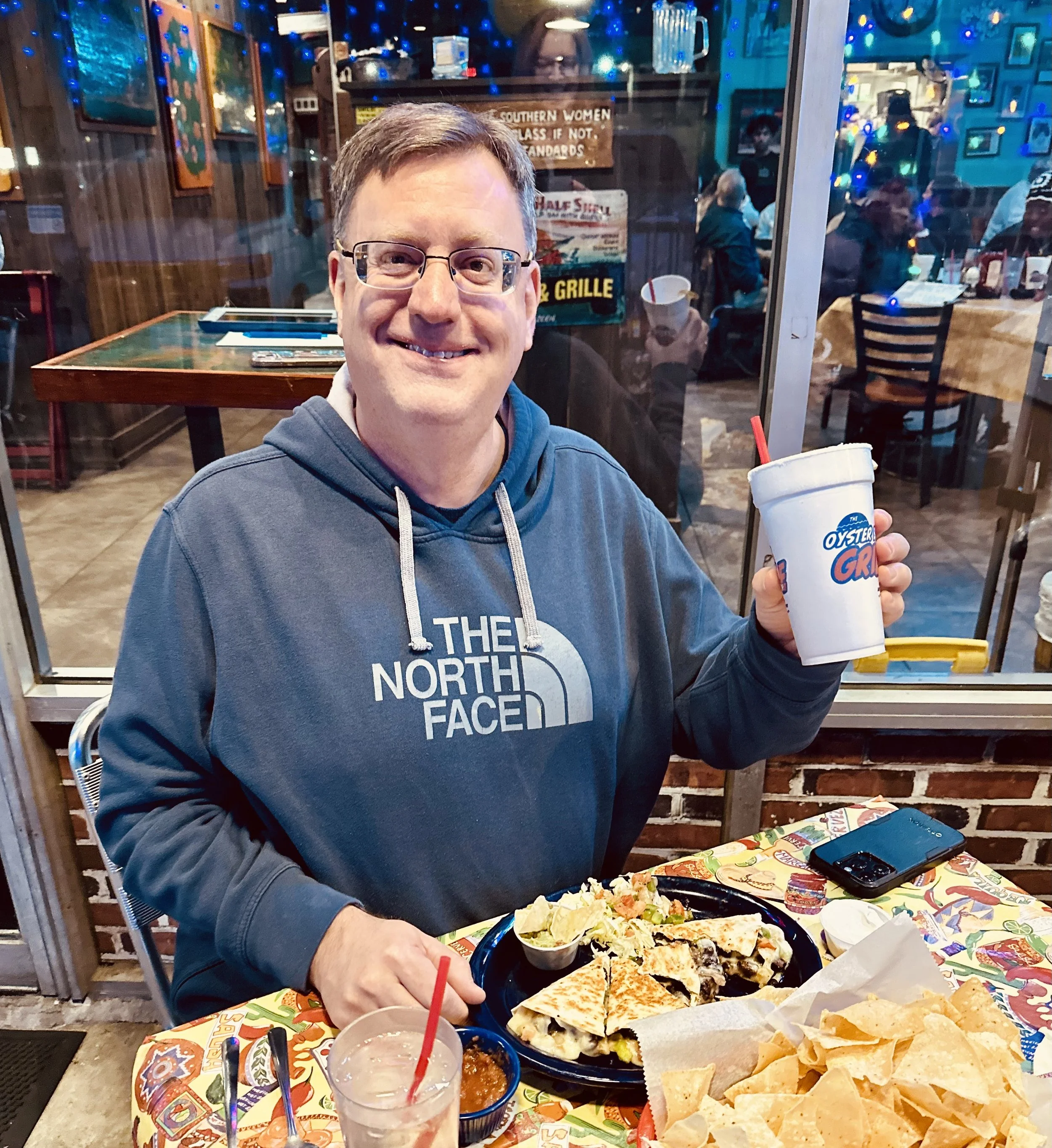 A man with glasses wearing a blue North Face hoodie sitting at a festive table with Mexican food including nachos, tacos, salsa, and drinks inside a restaurant with colorful decorations and artwork on the walls.