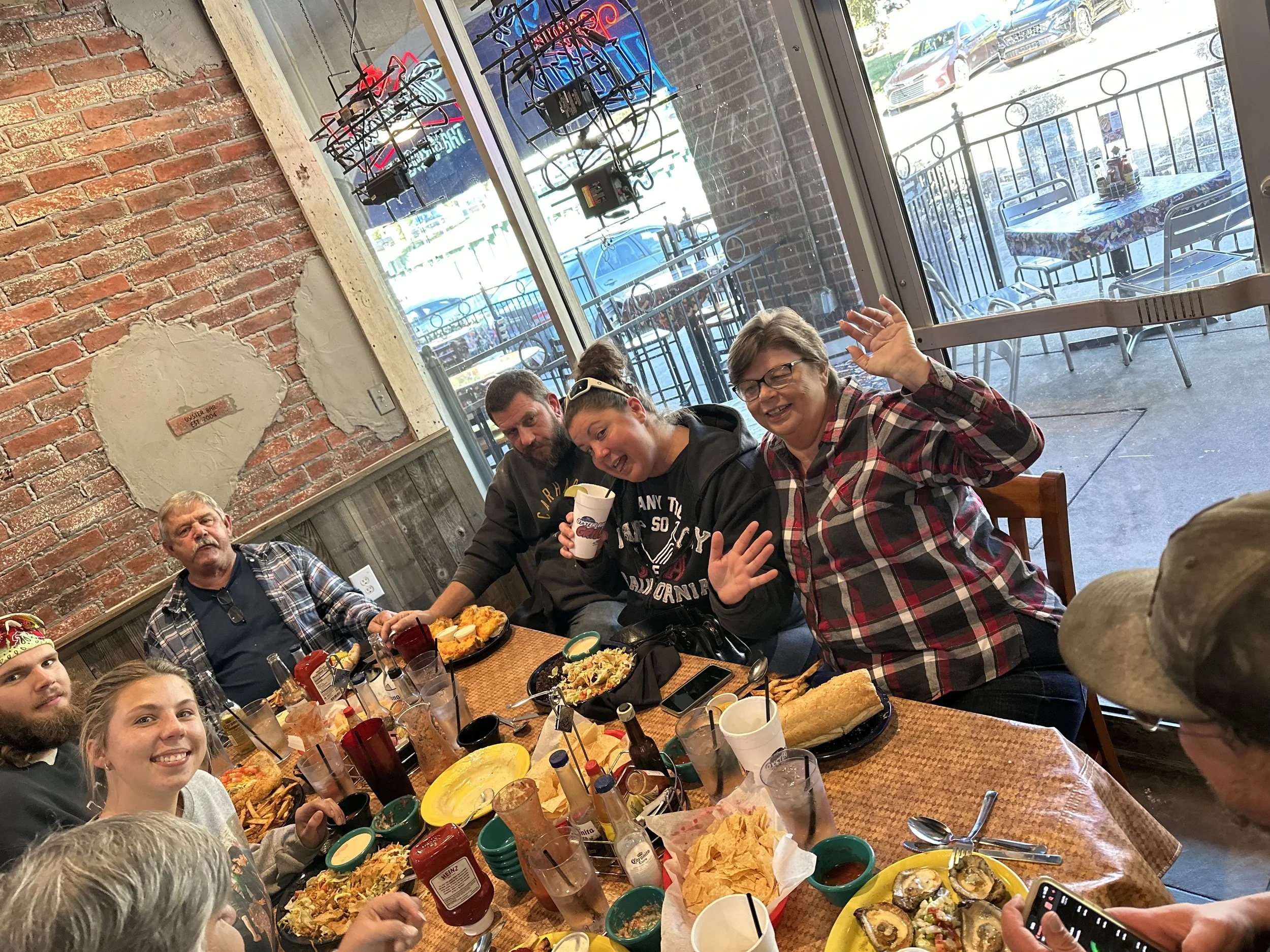 A group of people enjoying a meal at a restaurant, sitting around a table filled with various dishes like fries, sushi, and dips, inside a restaurant with exposed brick walls and large windows showing parked cars outside.