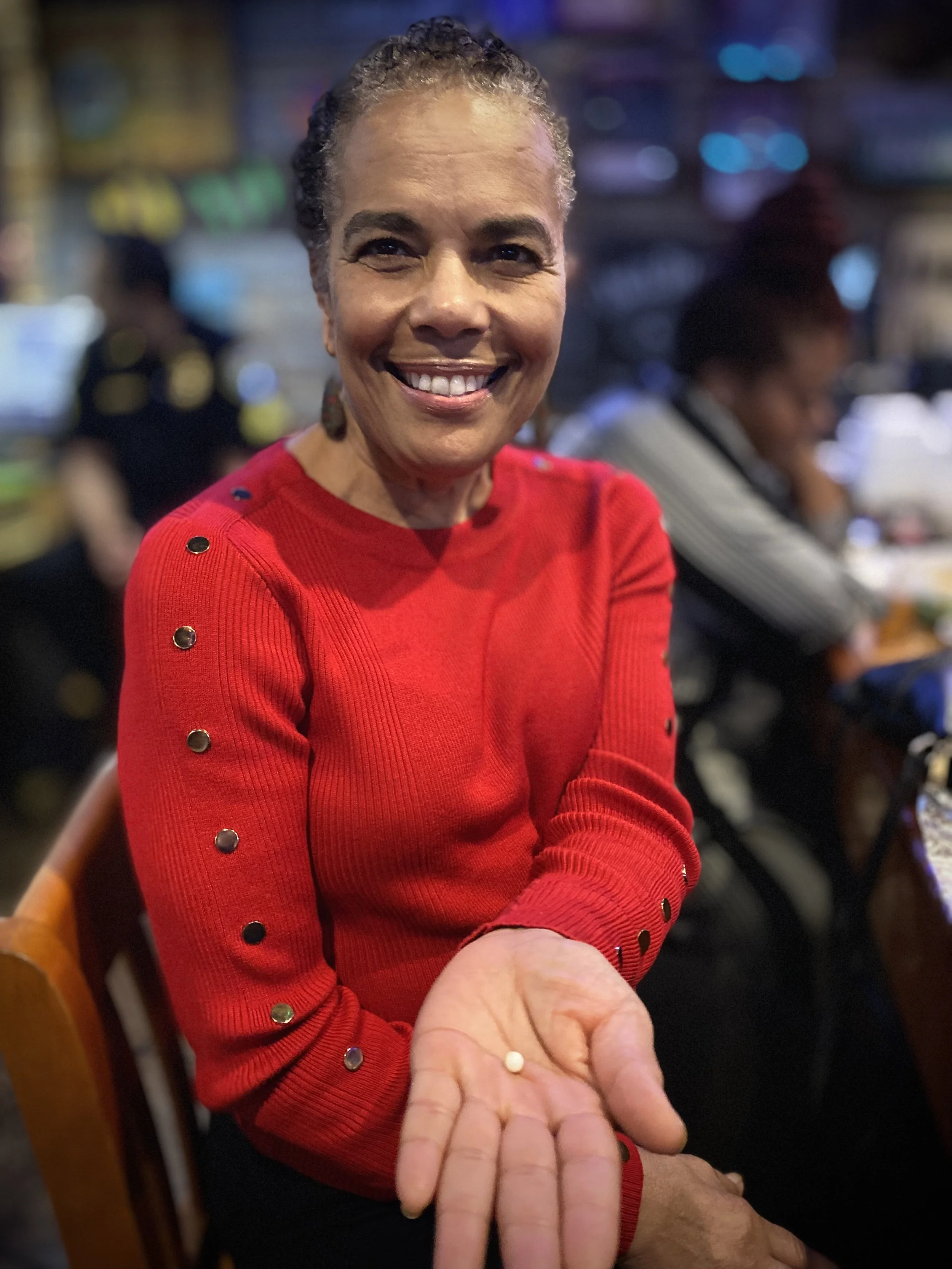 A woman in a red sweater with black buttons smiles as she holds a small pill in her palm, with a blurred background of a busy indoor setting.