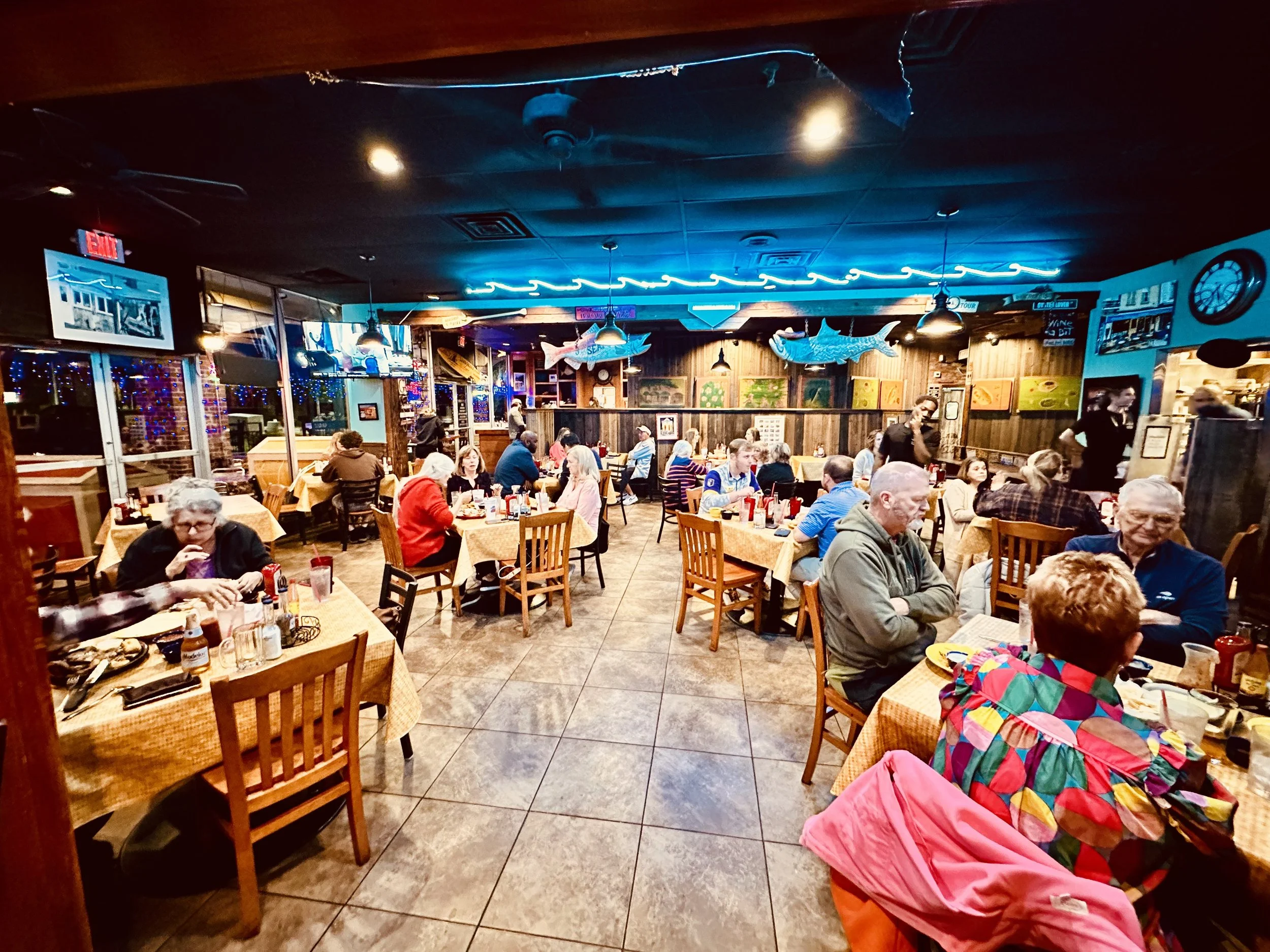 A busy seafood restaurant with people dining at tables covered with yellow checkered tablecloths, decorated with condiments and drinks. The interior features wooden walls, hanging fish decorations, neon blue lighting, and multiple televisions and art