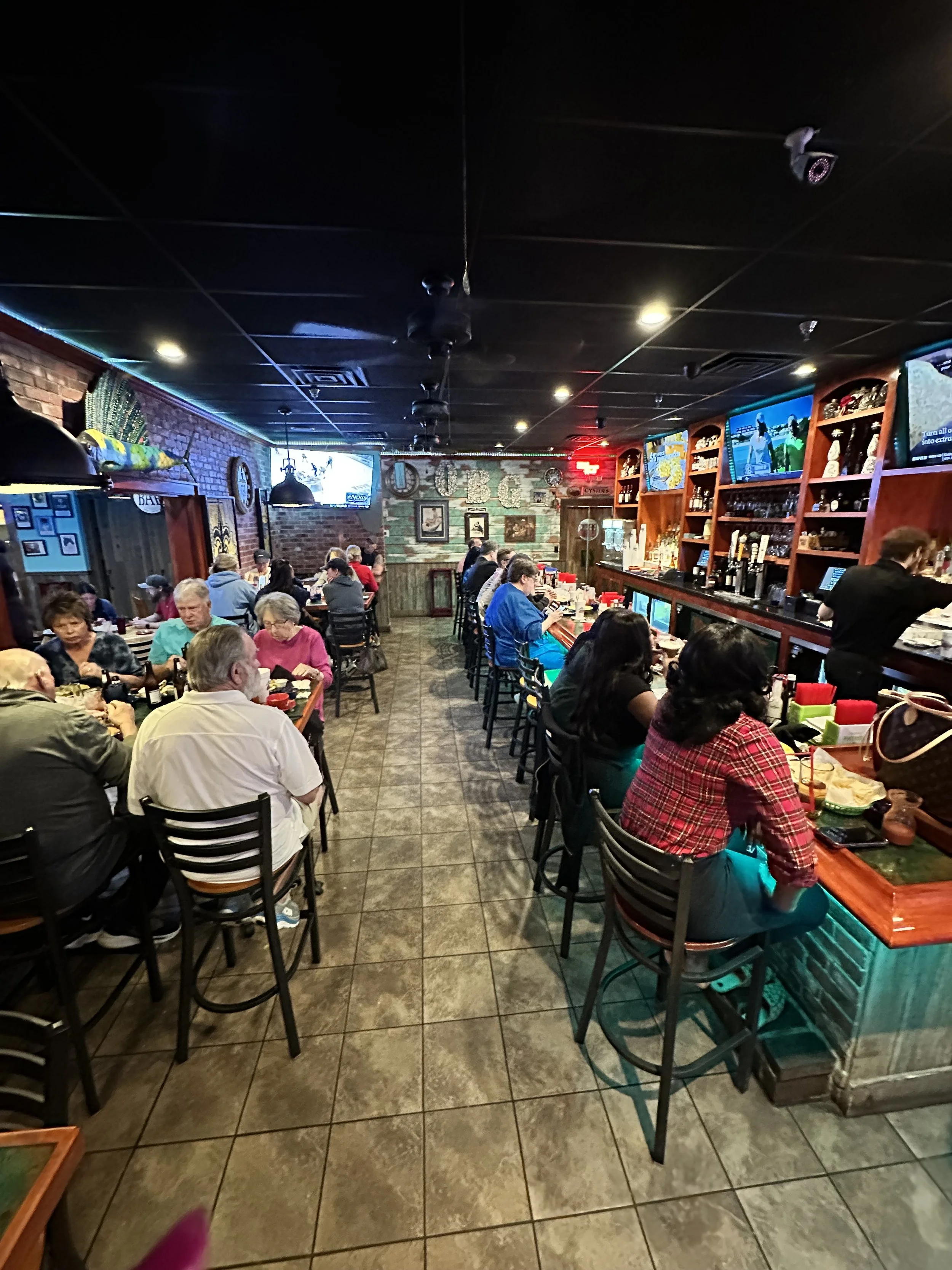 A crowded restaurant with patrons sitting at a long bar counter and tables, brick walls, and flat-screen TVs on the wall.