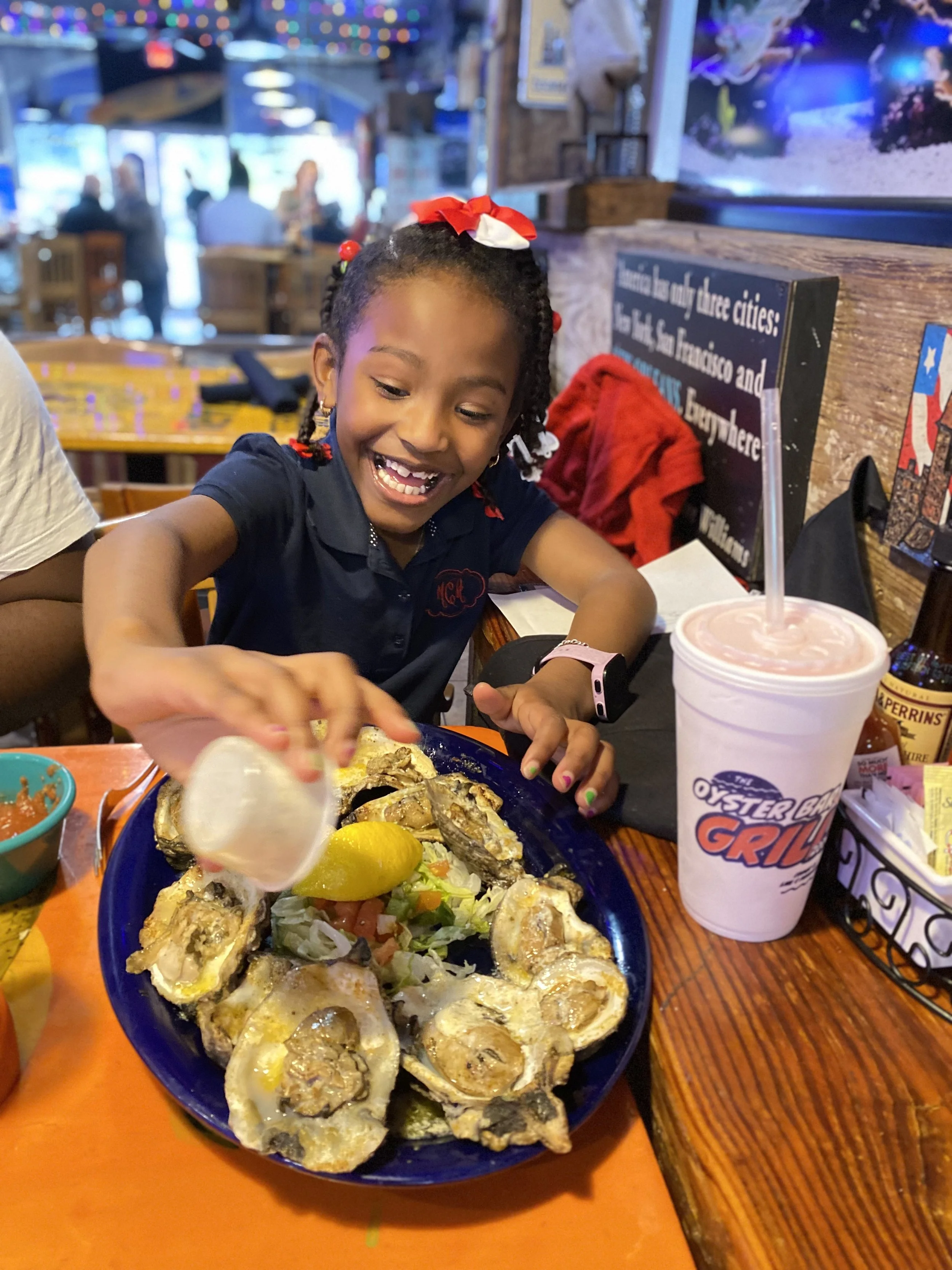 A young girl with braided hair, wearing a navy blue shirt and a red bow, smiling and reaching for food at a restaurant. In front of her is a large blue plate of oysters on a bed of lettuce and vegetables, with a lemon wedge. To her right, there is a 