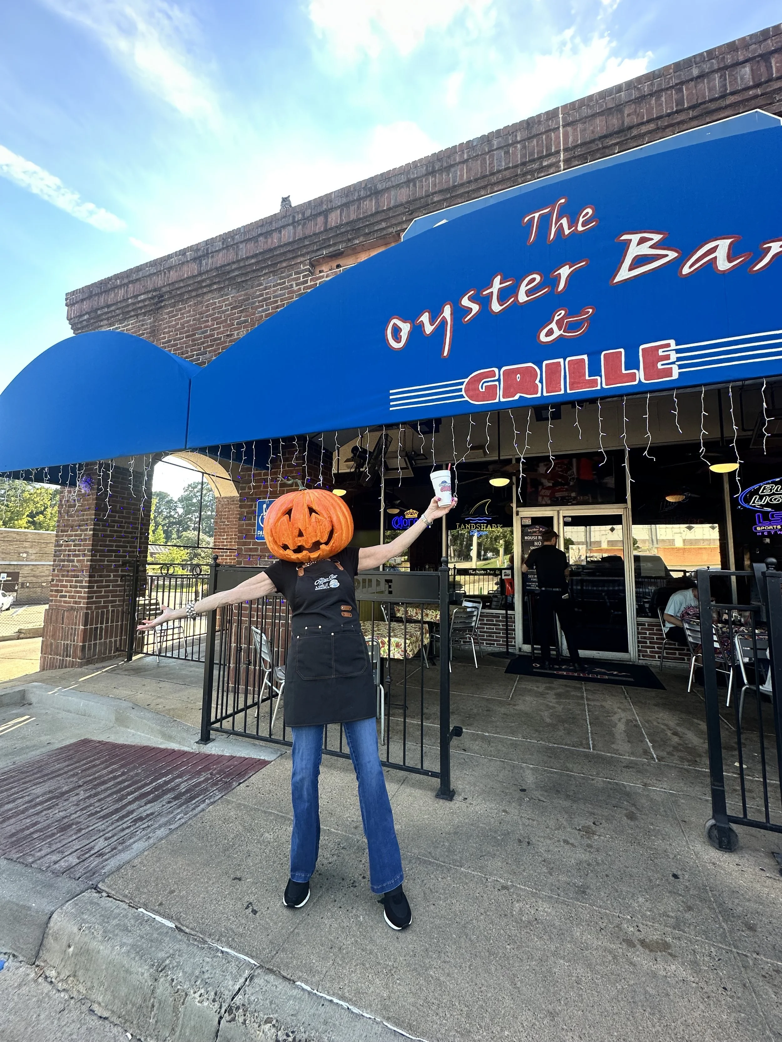 Person with a pumpkin head costume raising a drink in front of a restaurant with a blue awning that reads 'The Oyster Bar & Grille."