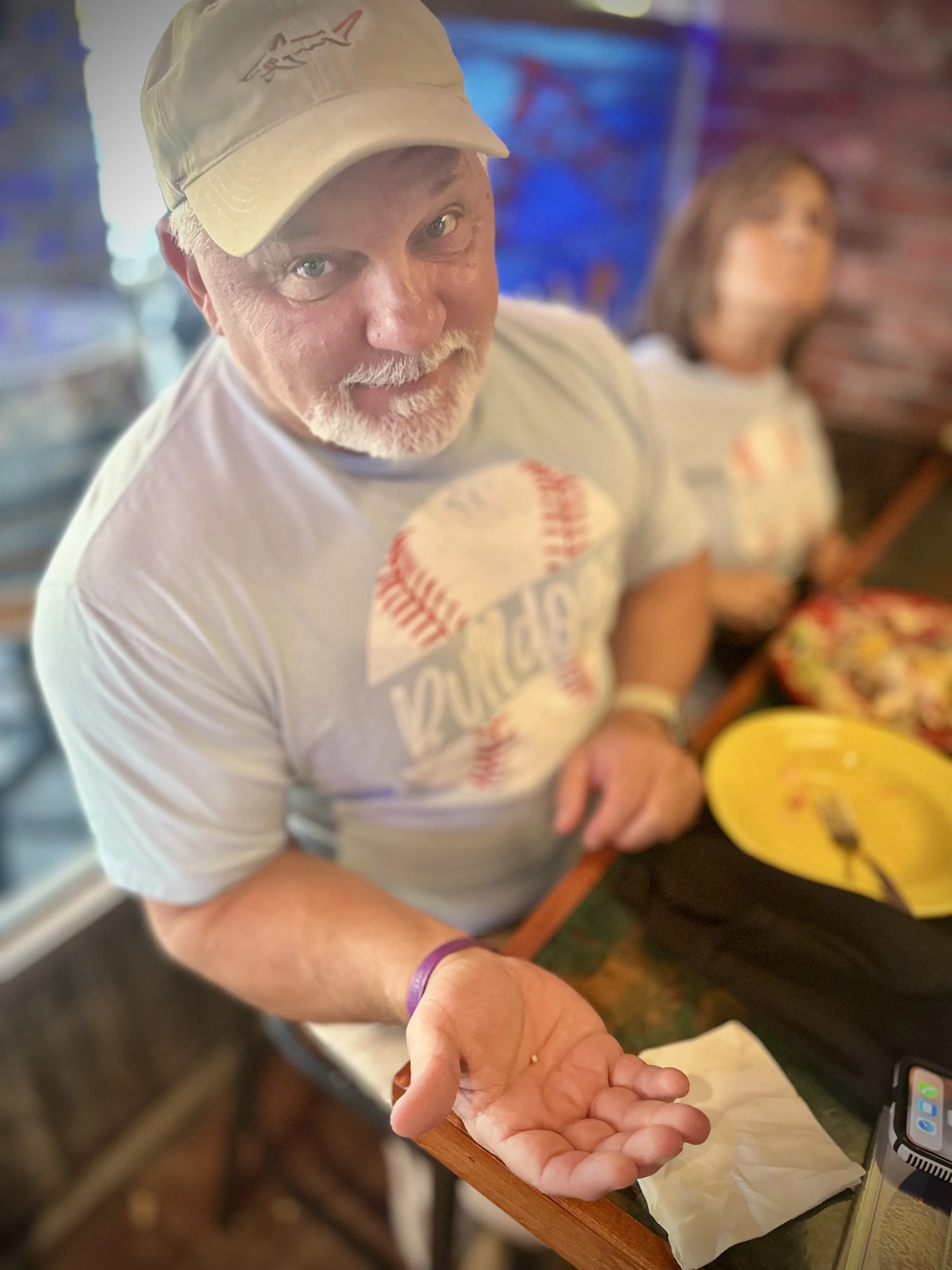 An elderly man with glasses, a light-colored cap, and a white T-shirt with red and blue writing, extending his hand toward the camera while standing at a counter with a yellow bowl, showing the pearl he found in his oyster