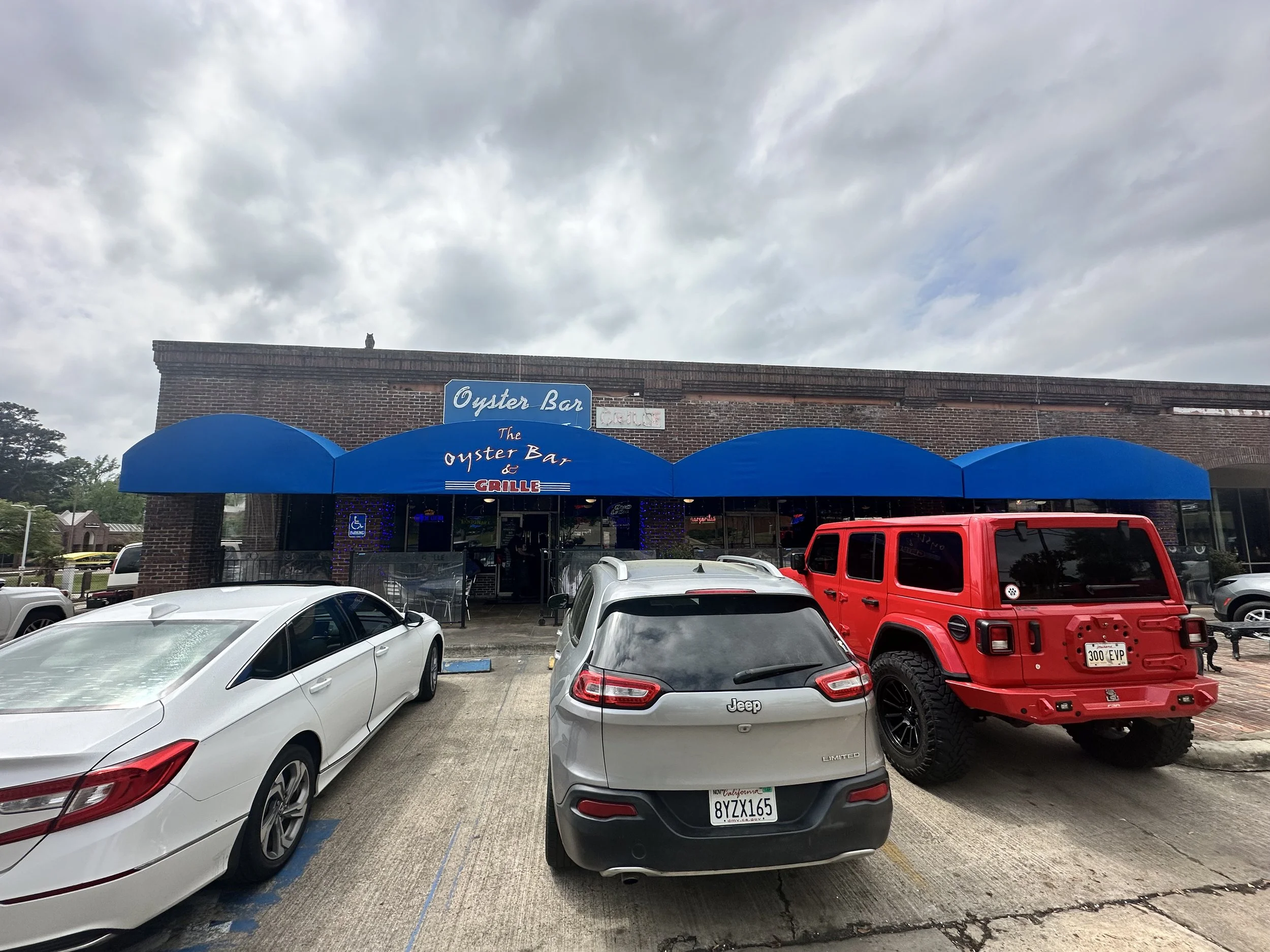 Parking lot scene in front of a building with a blue awning labeled 'Oyster Bar'. Several cars are parked, including a white car, a silver Jeep, and a red vehicle. A cat is sitting on the roof of the building under a cloudy sky.