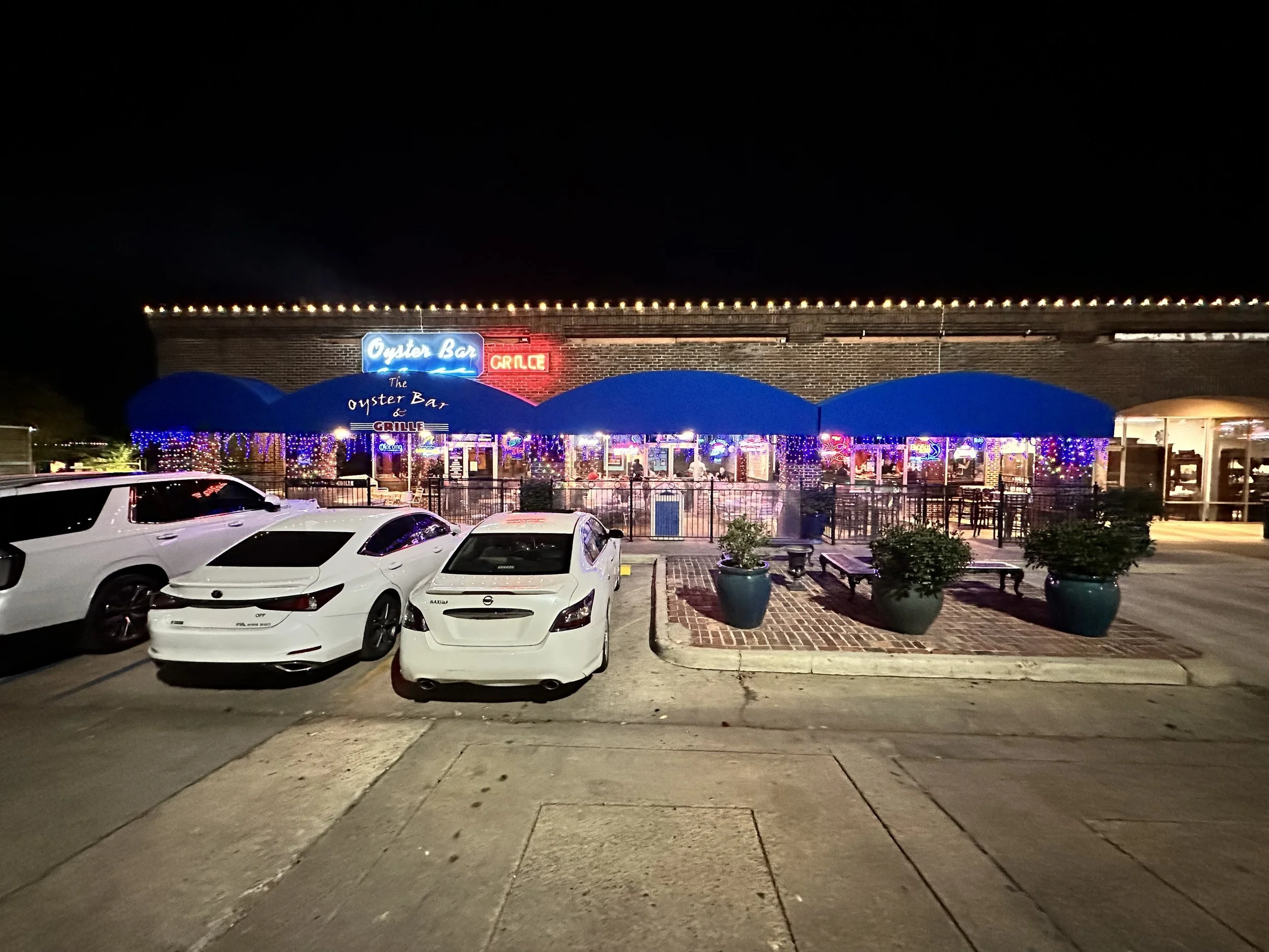 Nighttime scene of a restaurant named Oyster Bar and Grille with neon signs, string lights, and a parking lot with several white cars in the foreground.