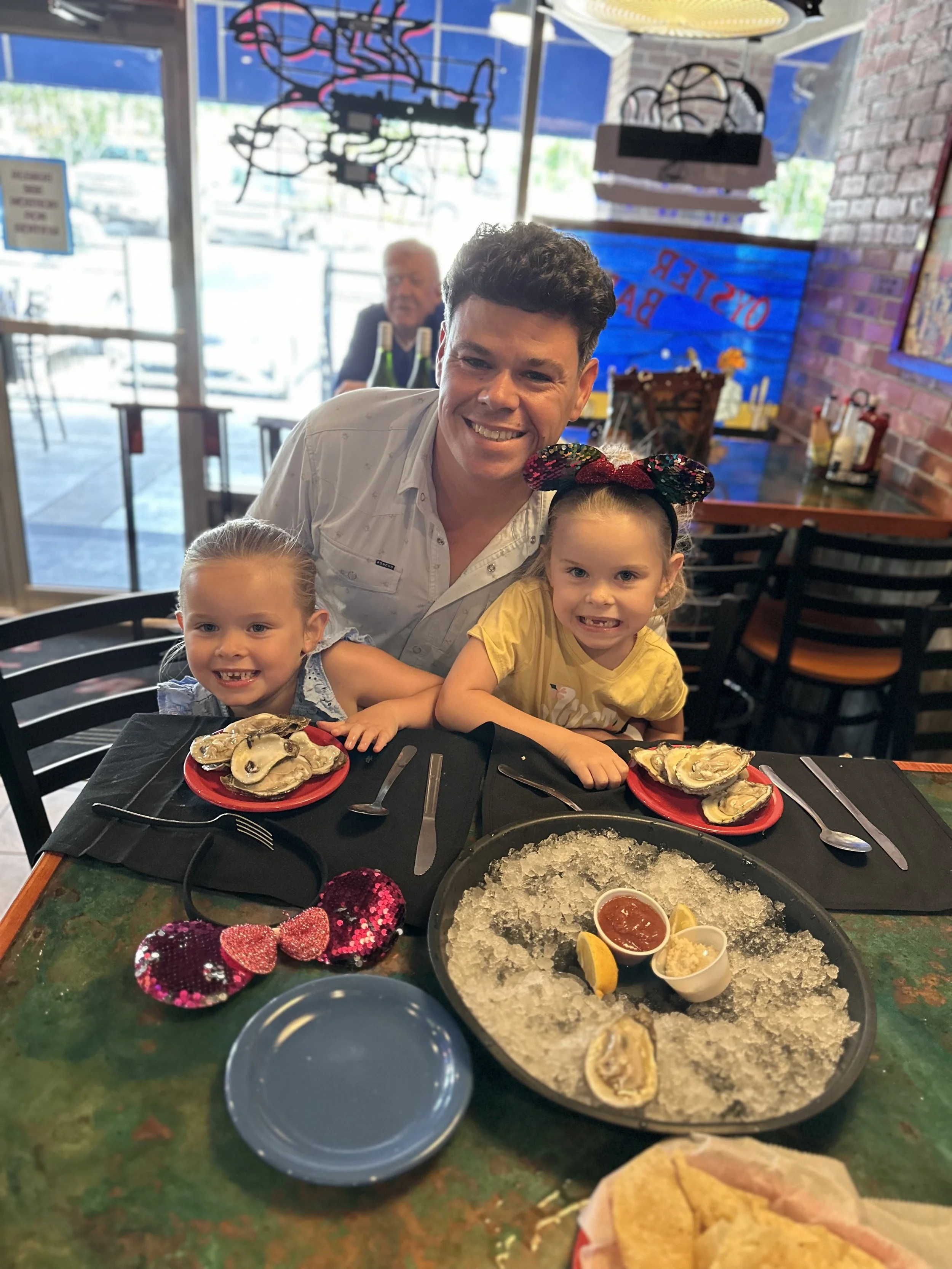 Man and two young girls smiling at a table with plates of oysters, lemon wedges, and condiments in a restaurant with brick walls and colorful neon signs in the background.