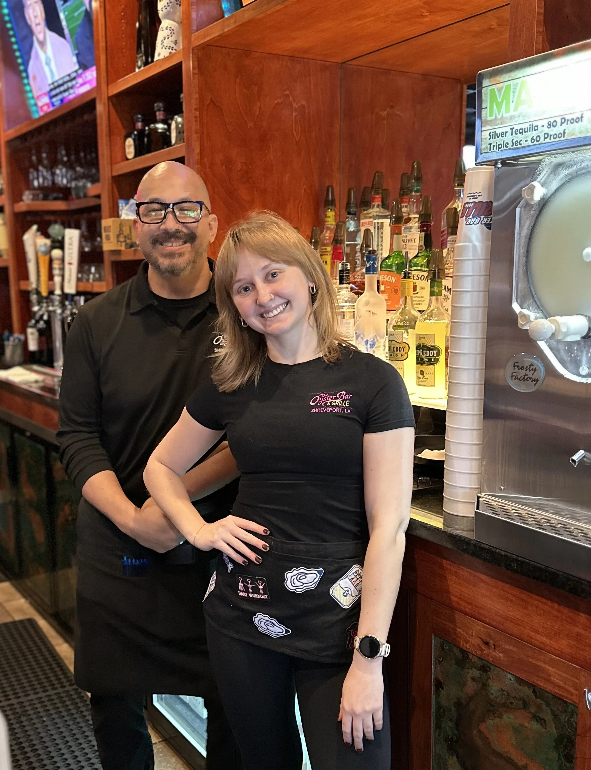 Two waitstaff, a bald man with glasses and a woman with shoulder-length hair, smiling and posing behind a bar with alcohol bottles in the background.