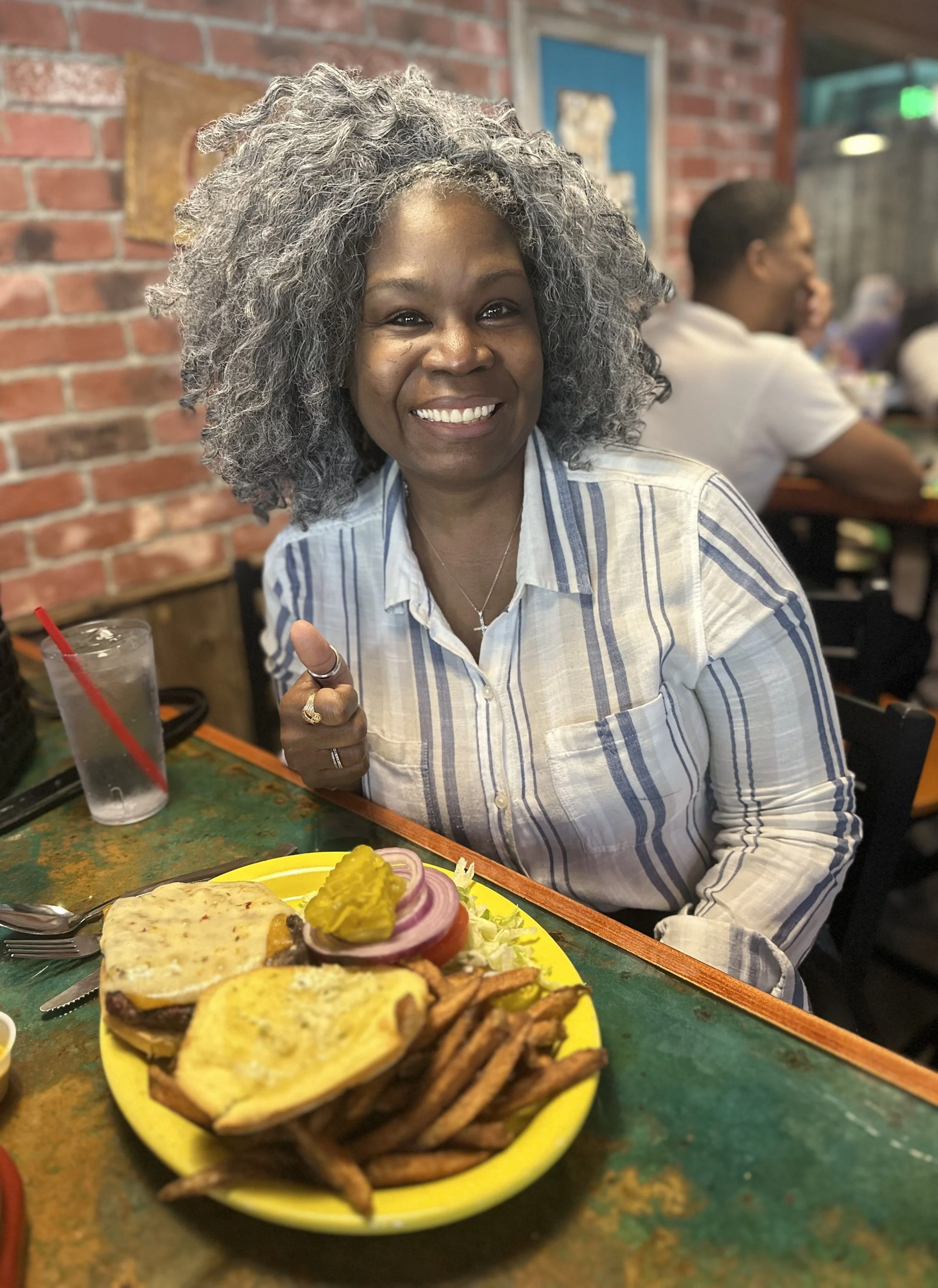 A smiling woman with gray curly hair wearing a striped button-up shirt sitting at a restaurant table with a yellow plate of food in front of her. The plate contains various foods including jalapenos, slices of onion, and grilled items. In the backgro