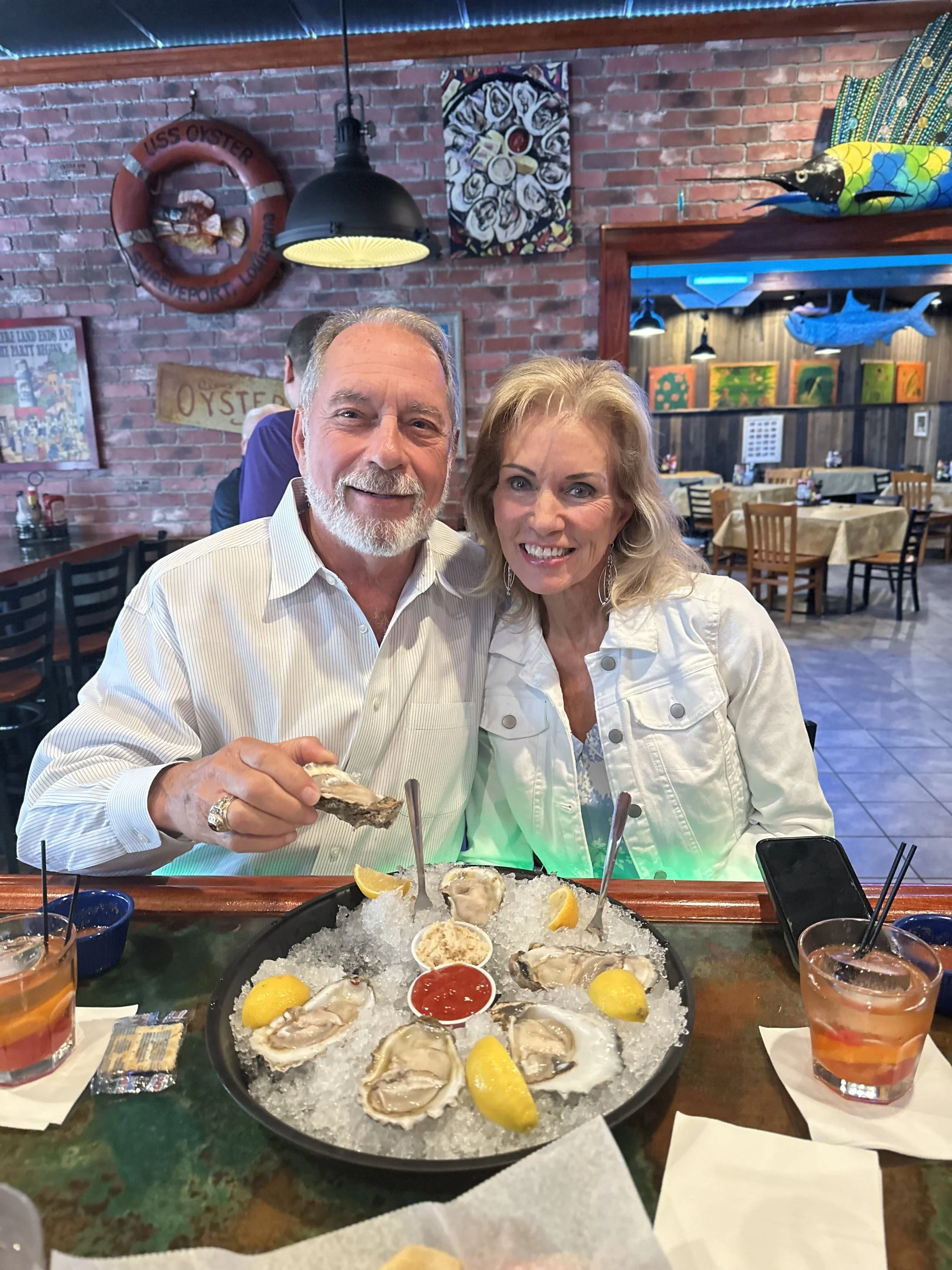A man and woman sitting at a restaurant table with a large platter of fresh oysters on ice, garnished with lemon wedges and sauces, surrounded by glasses of drinks. The background features a brick wall with marine-themed decorations.