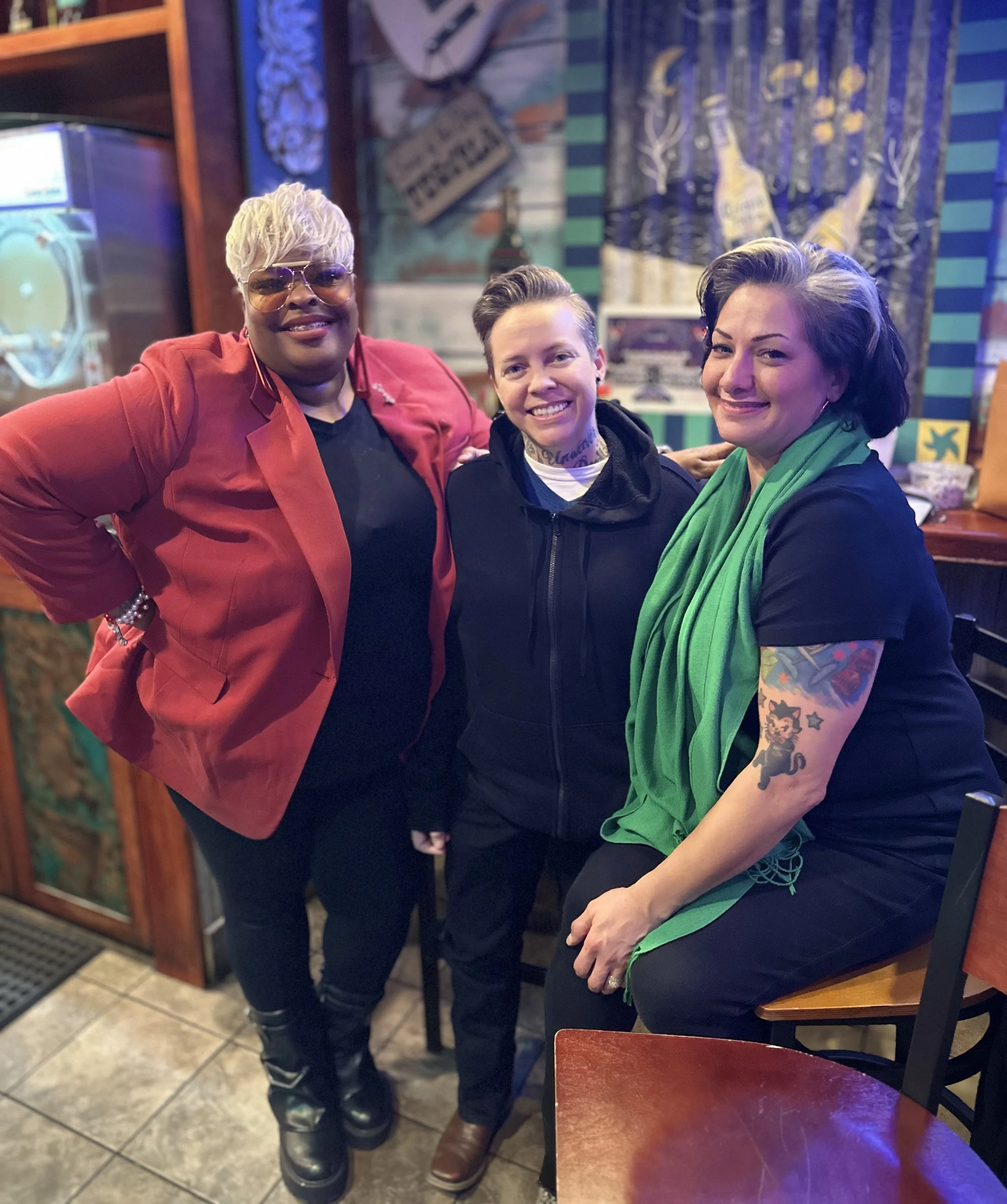 Three women standing close together inside a restaurant, smiling at the camera. The woman on the left has short, platinum blonde hair, is wearing sunglasses and a red blazer. The woman in the middle has short, light brown hair and tattoos on her neck