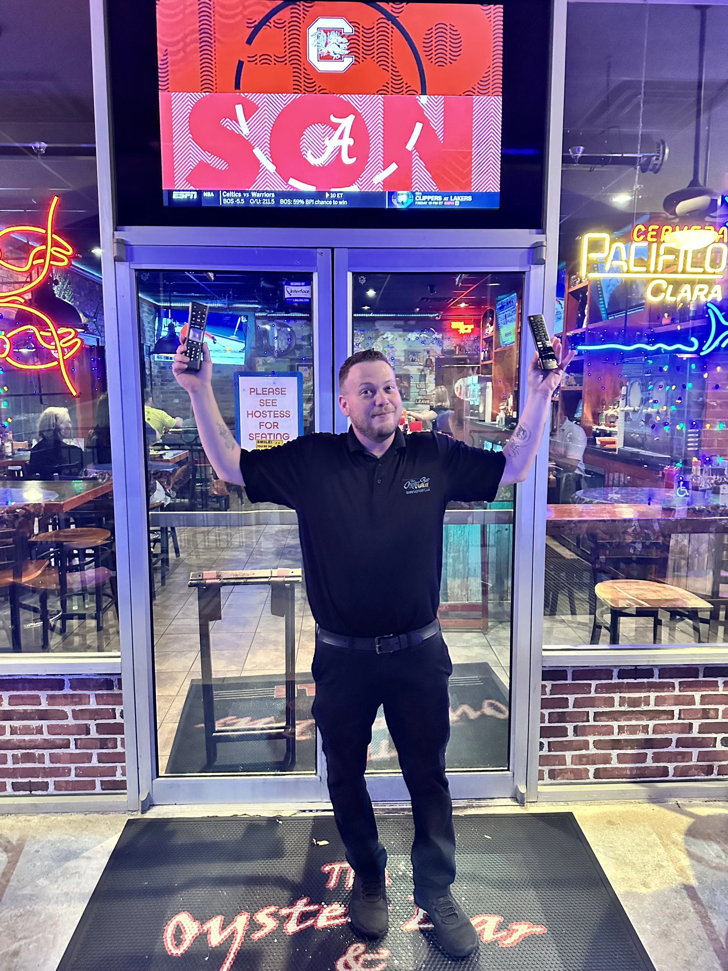 A man in a black uniform standing inside a restaurant, holding a remote control in each hand, with a welcoming smile. The restaurant has brick walls, neon signs, and a glass door entrance. There is a sign on the door that says 'Please see hostess for