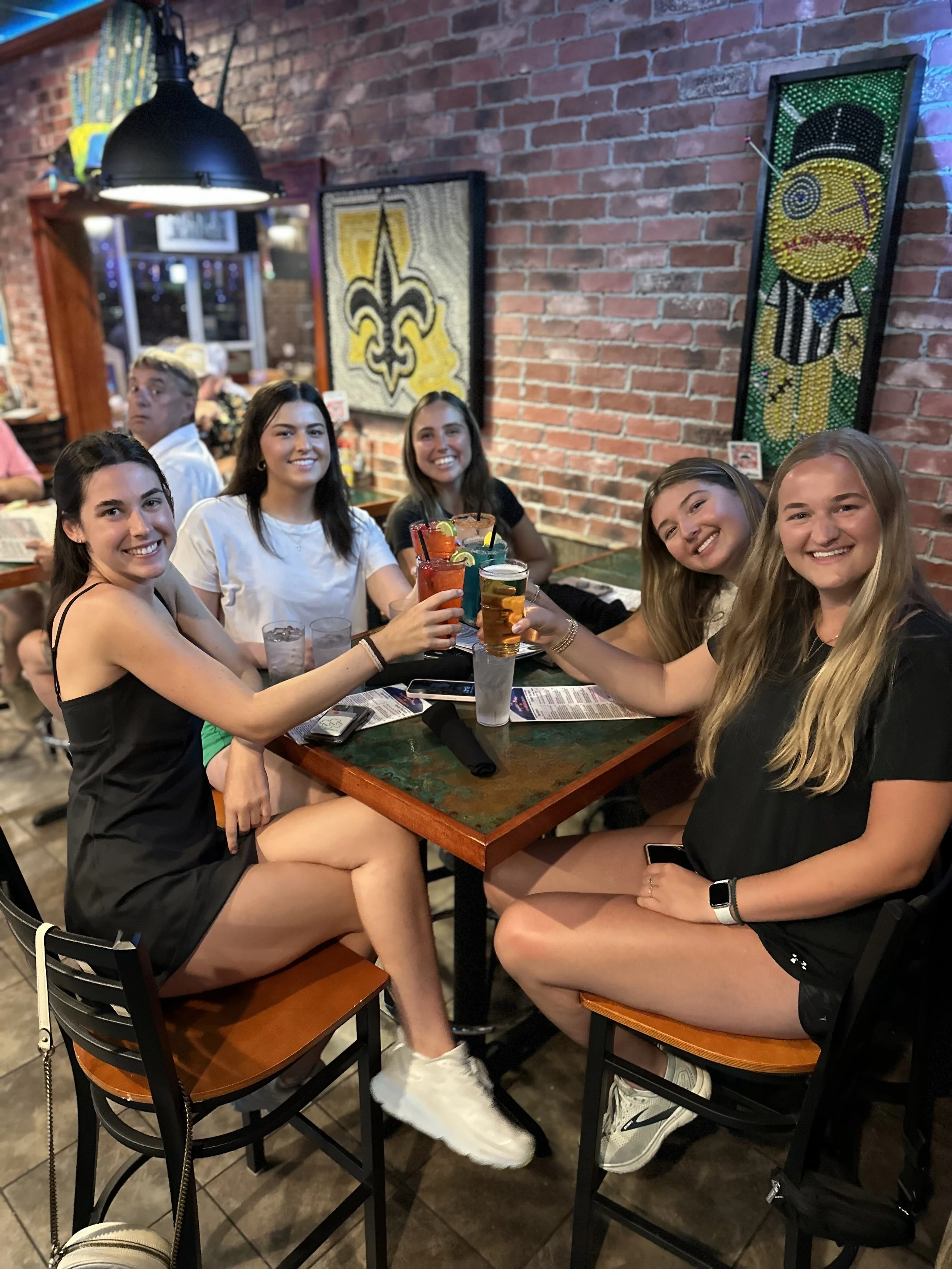 Five smiling women with drinks at a restaurant table with a brick wall and decorative artwork in the background.
